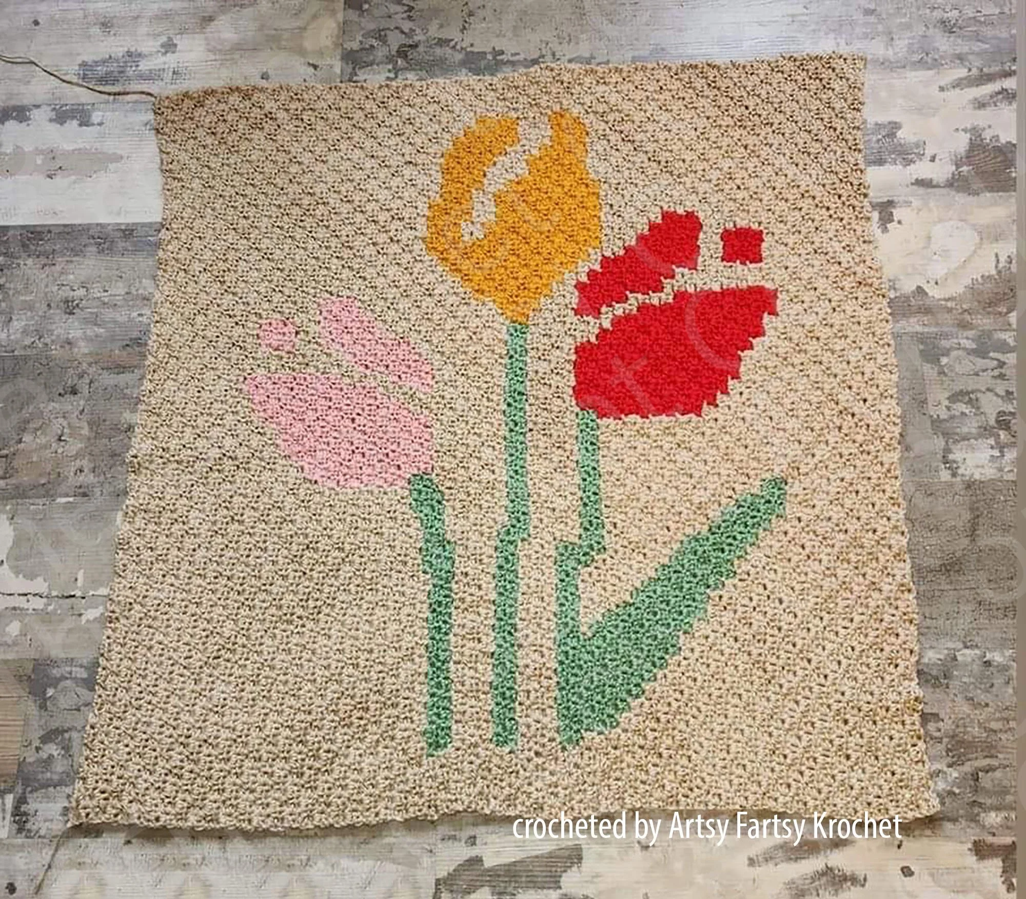 A small crochet afghan with a pixel-style design of three tulips in red, yellow, and pink, laid flat on a textured surface.