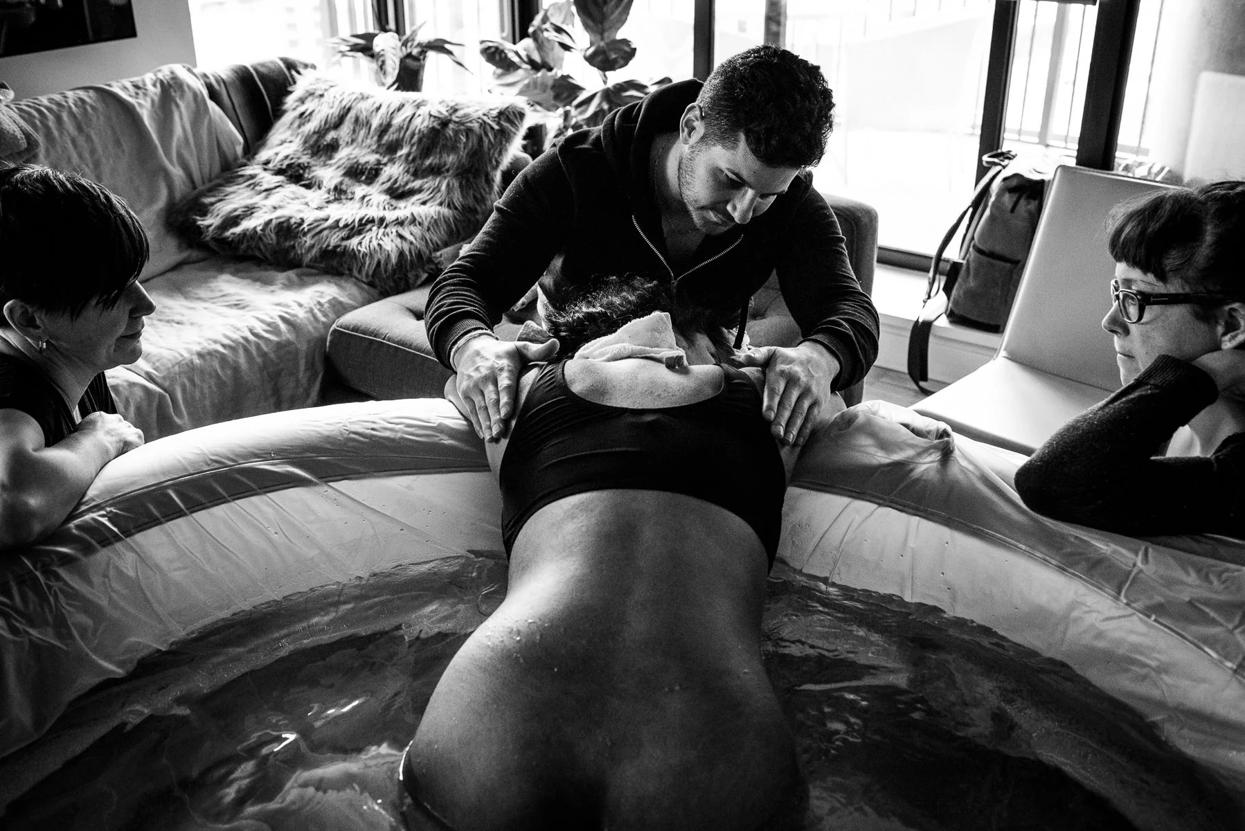 husband, midwife, and doula supporting a woman in labor while sitting in a birth pool at home.