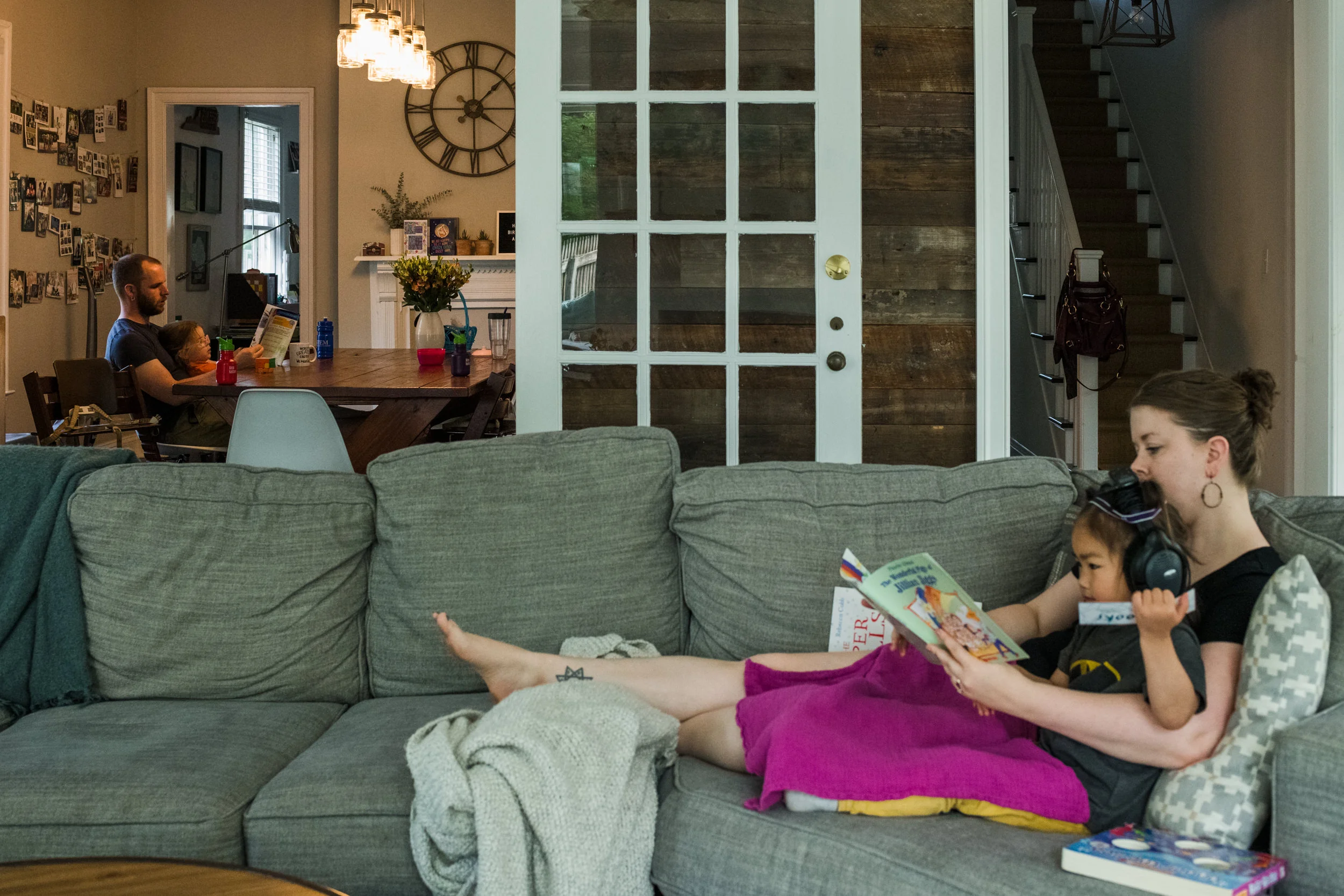 mom and daughter reading together on the couch while dad and other daughter reads together at the dining table.