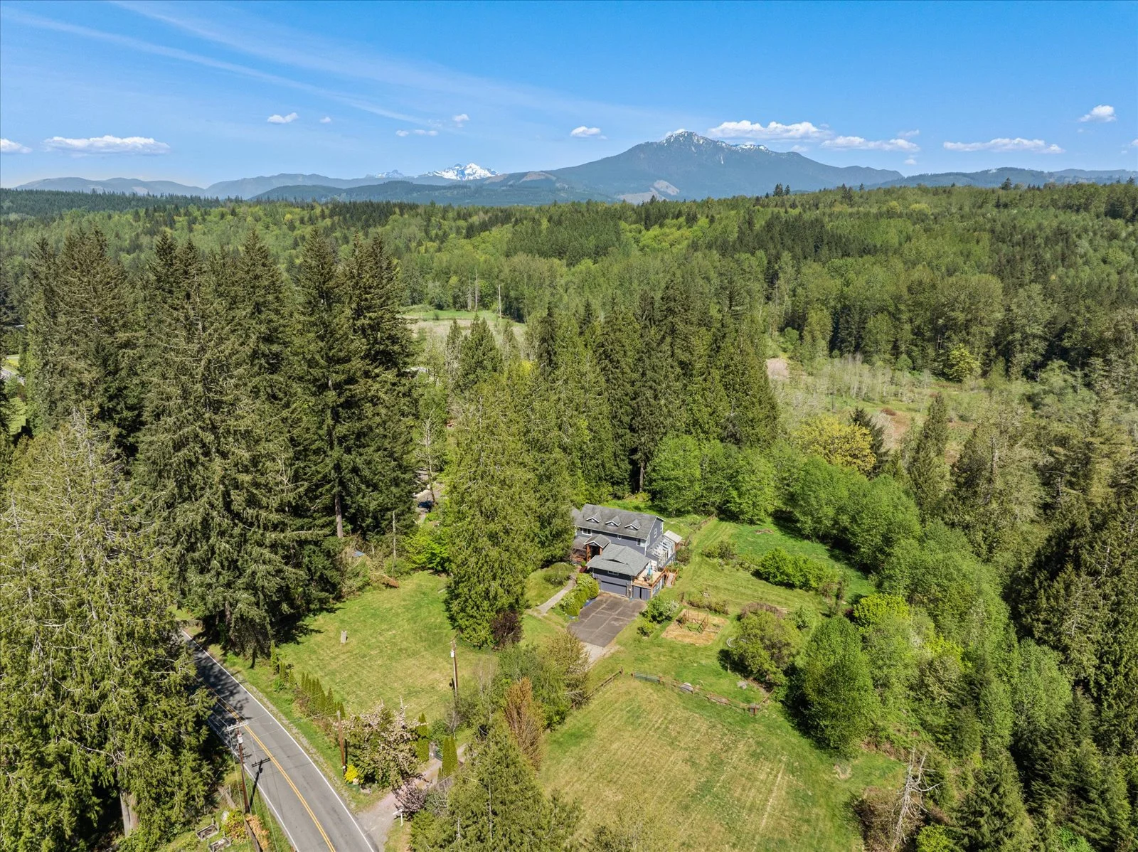Aerial view of a house surrounded by tall trees, with a winding road in the foreground and mountains in the background under a blue sky with a few clouds.