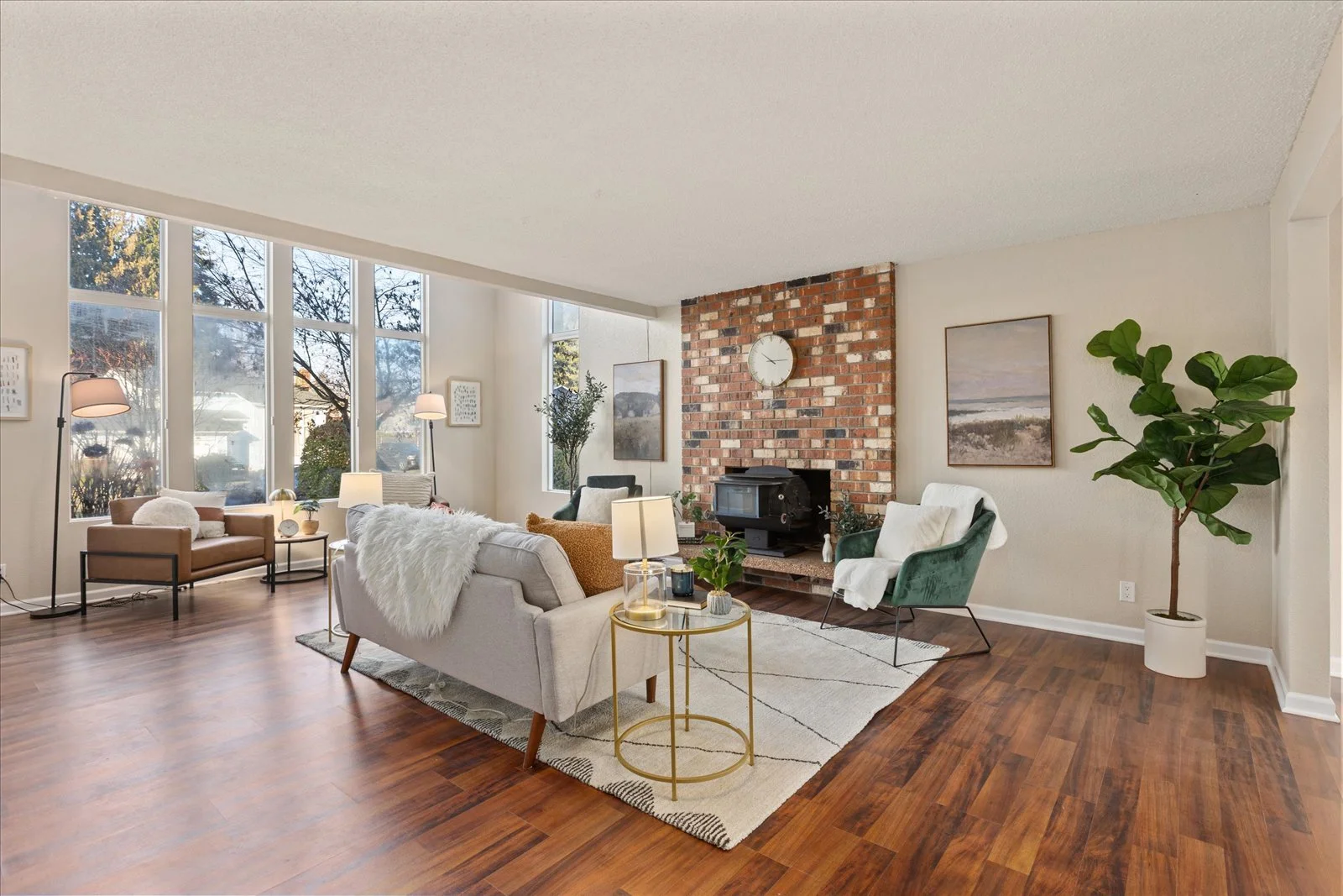 A spacious living room with large windows, a brick fireplace, a white sofa with cushions, a green armchair, floor lamps, and a potted plant on hardwood floors.