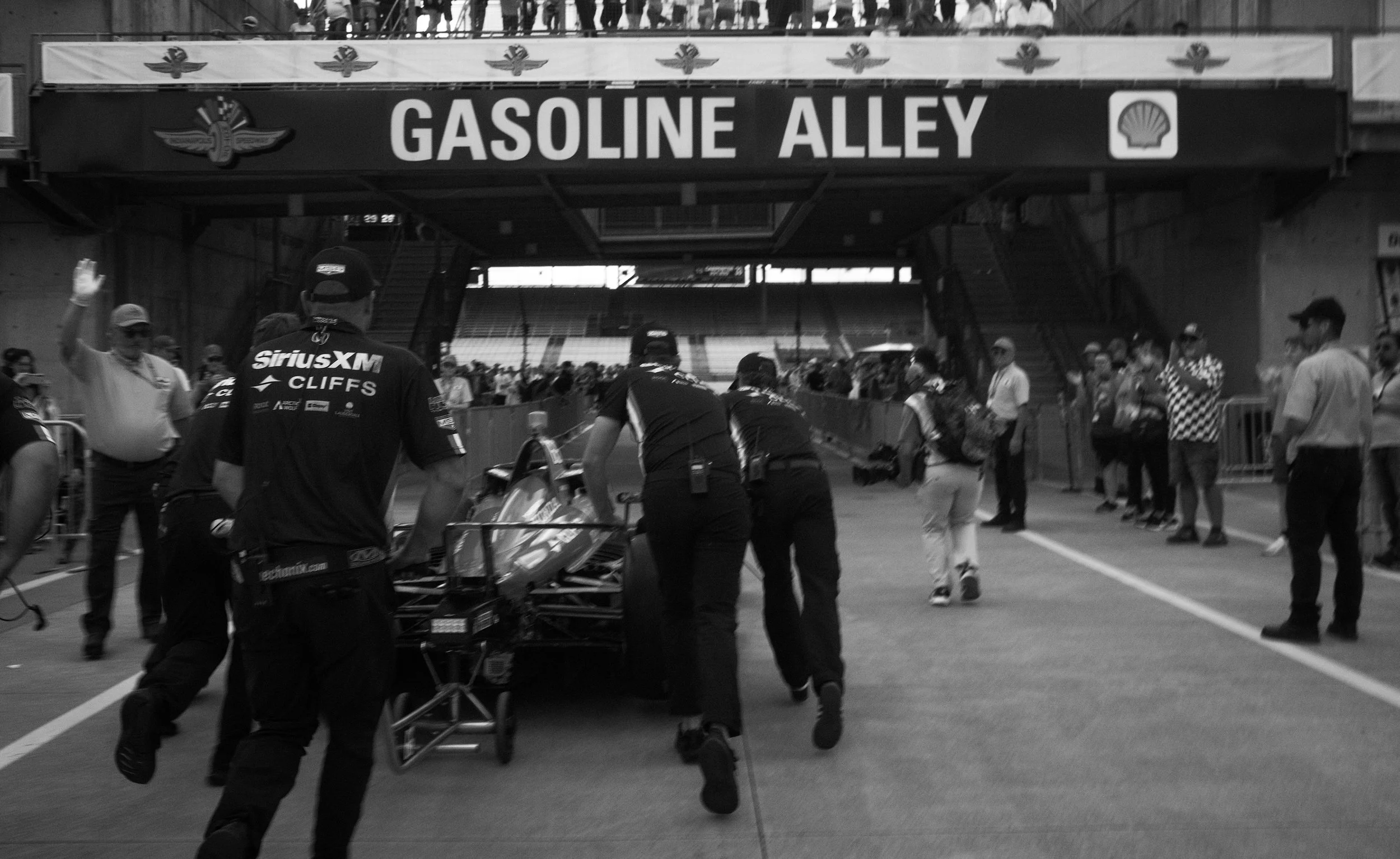 Race car team pushing car on track under a sign that reads 'Gasoline Alley' with people watching.