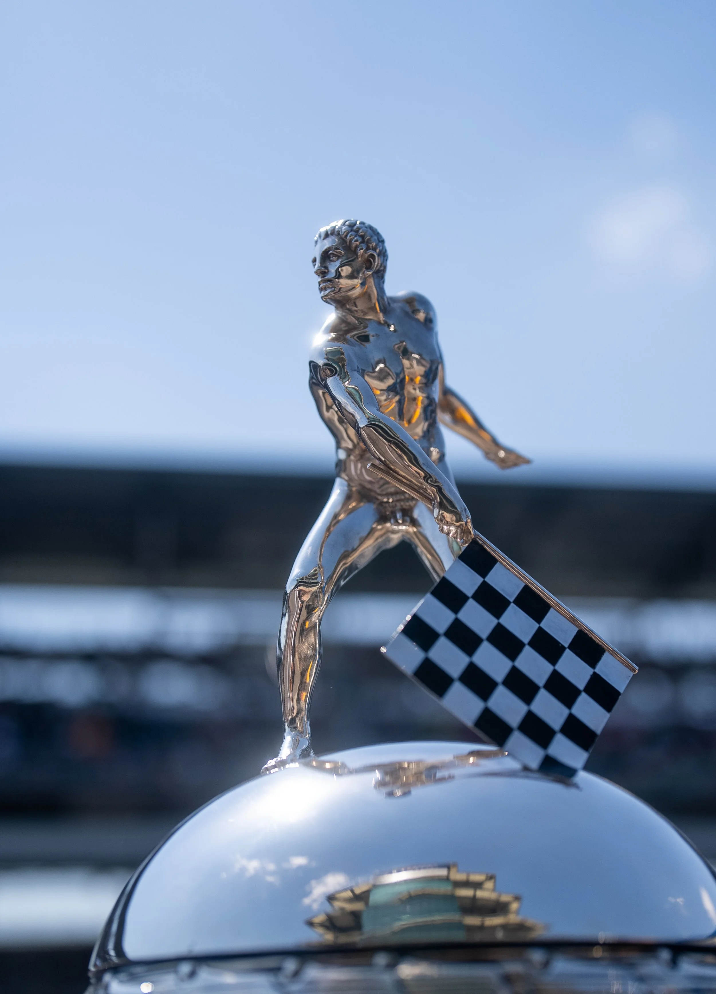 Close-up of a chrome trophy figure of a male race car driver holding a checkered flag, mounted on top of a shiny car hood with a blurred background and a partly cloudy sky.