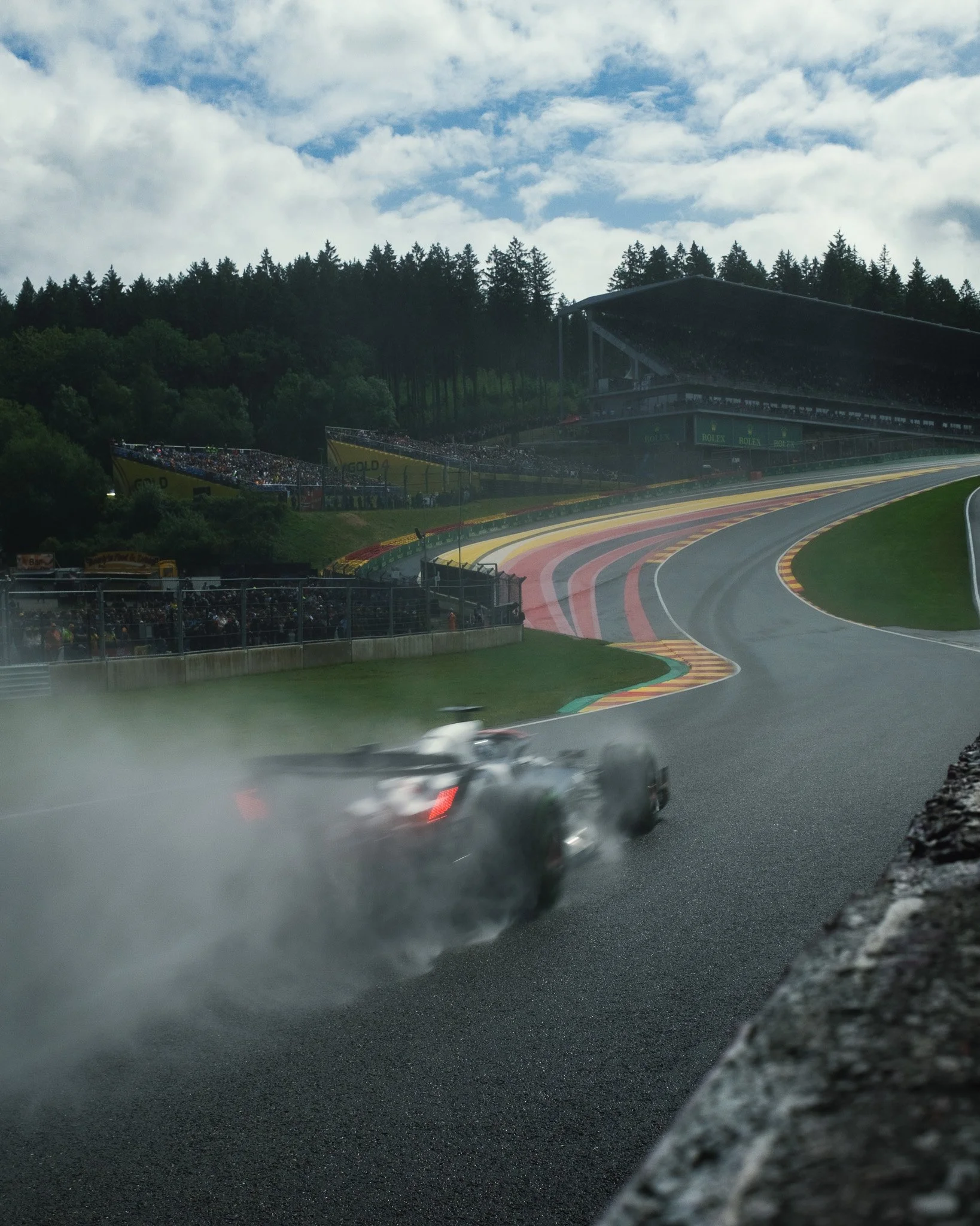 A race car taking a corner on a wet racetrack during a race, with a clouded sky and a grandstand filled with spectators in the background.