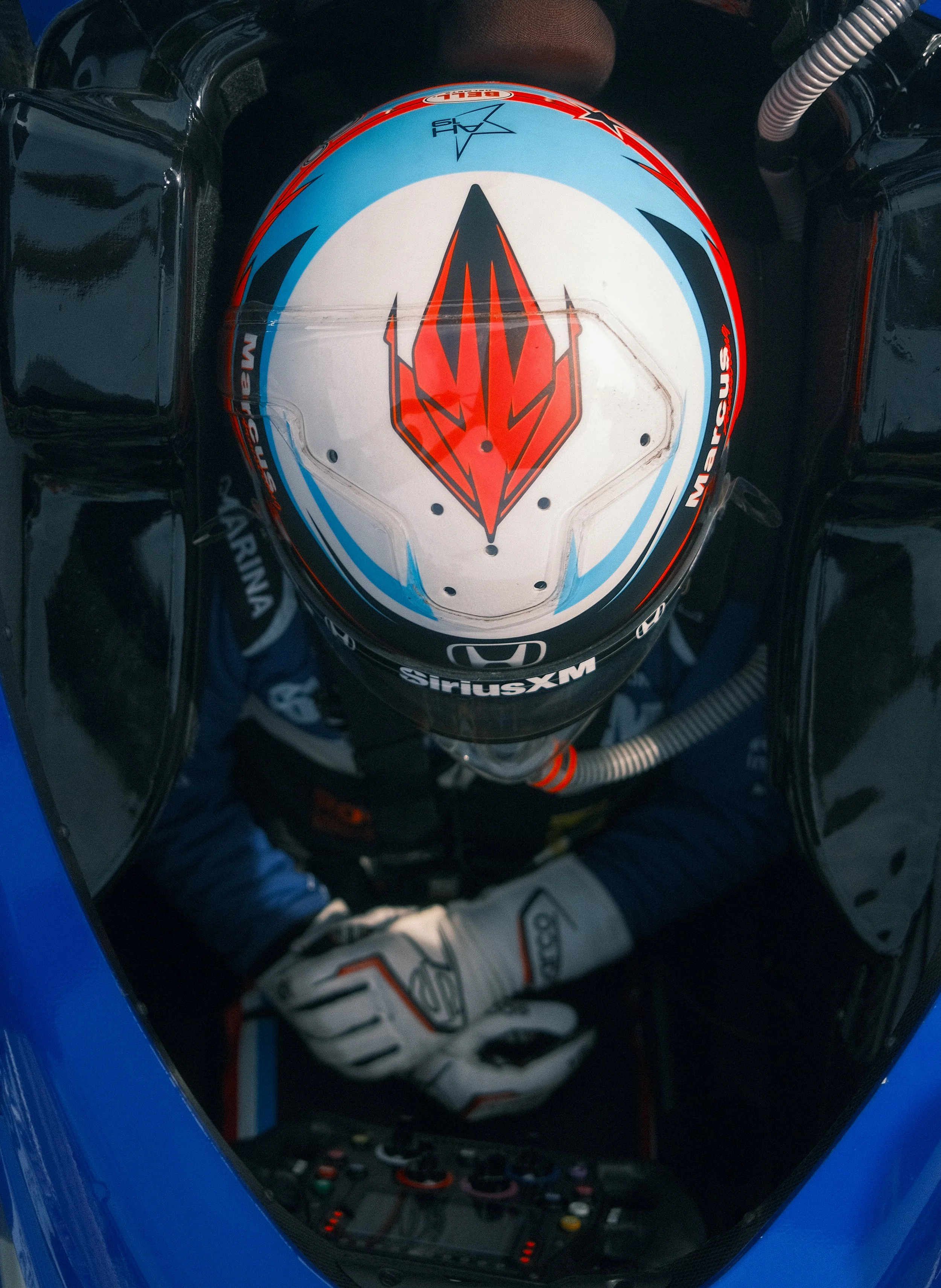 Top view of a race car driver wearing a helmet with a red and black design, sitting in a blue race car with digital controls and equipment visible in front of him.