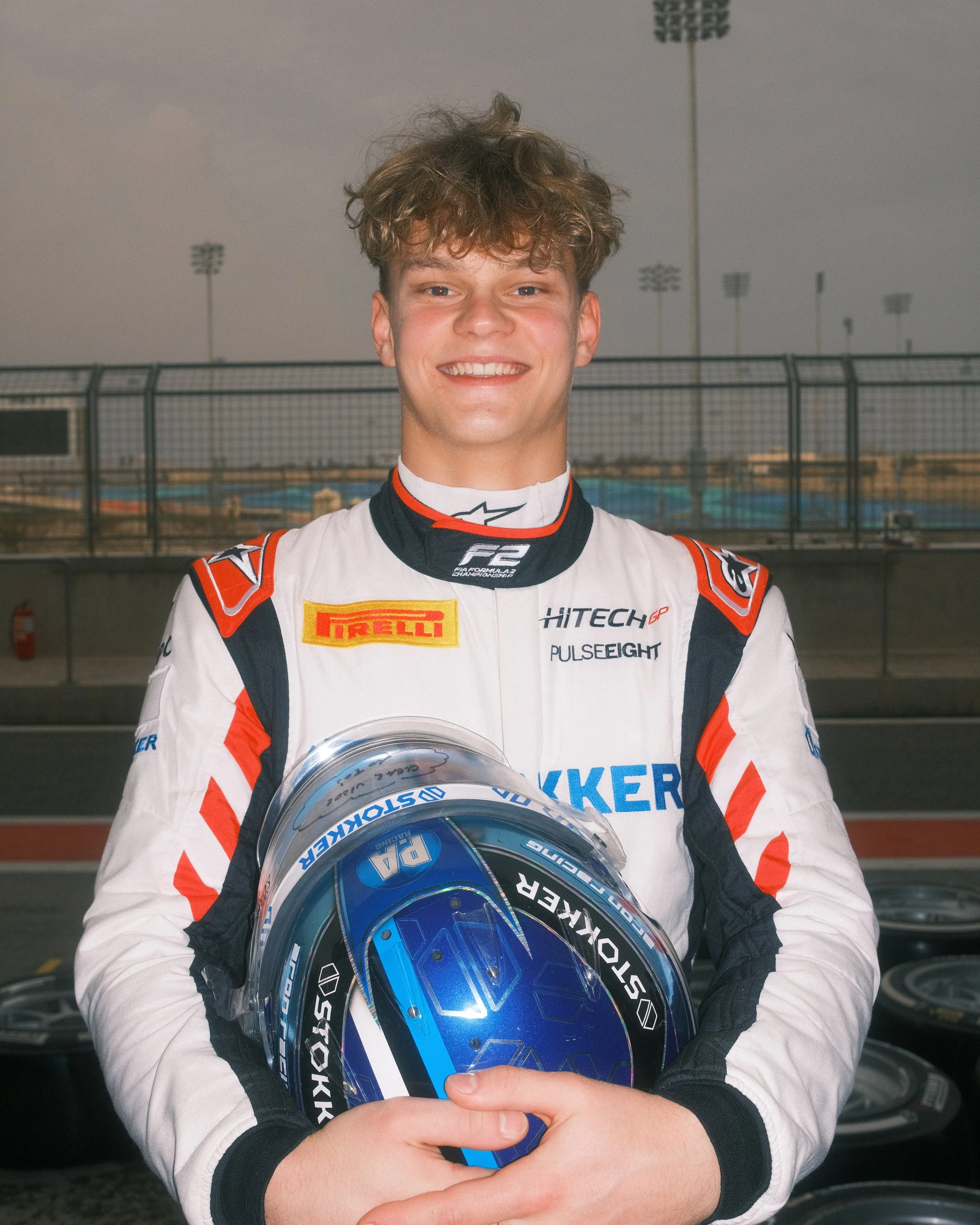 Race car driver Paul Aron in racing suit holding a blue helmet, standing on race track.