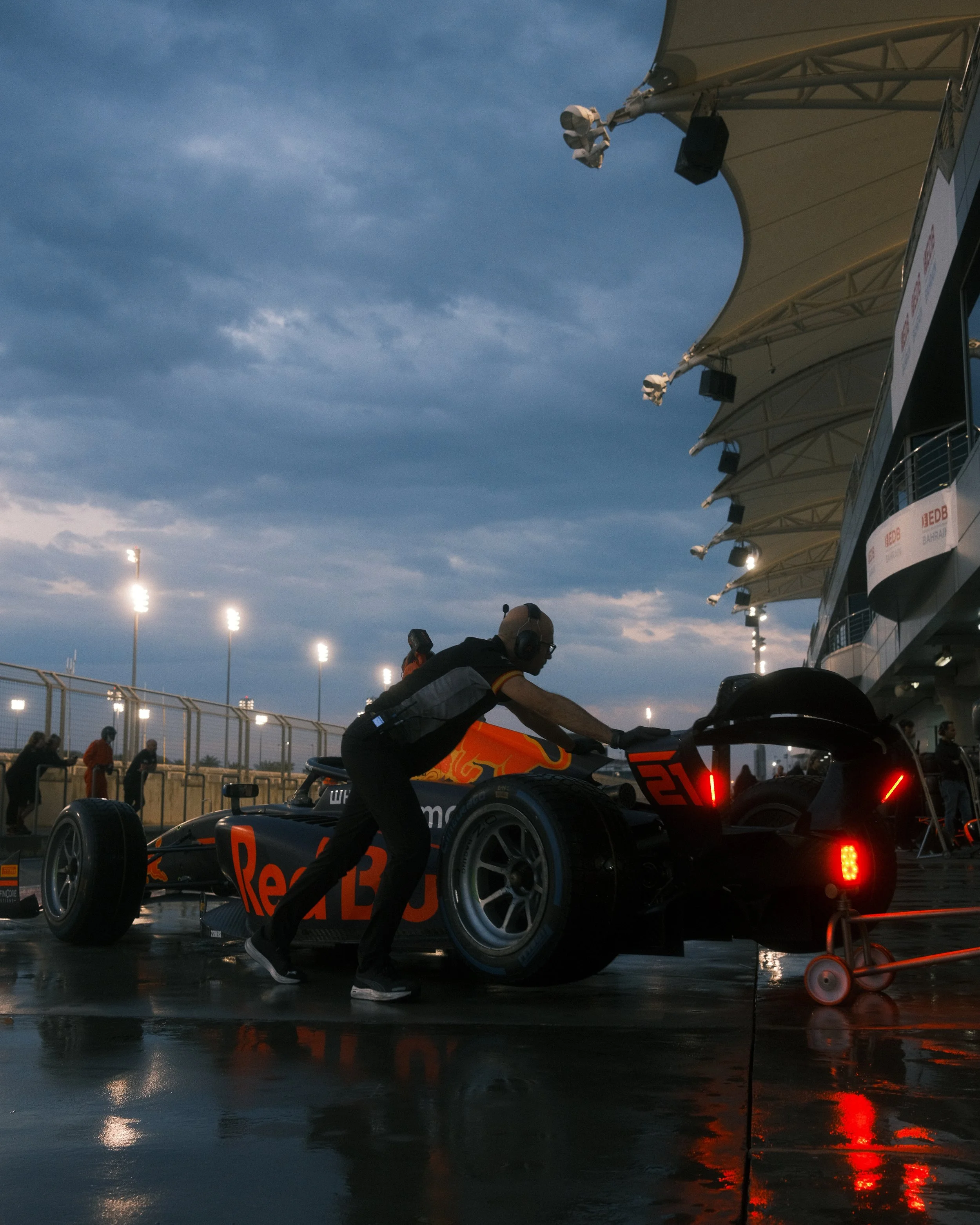 A Hitech racing team member in a pit stop area checks a black and orange Formula 1 race car with Red Bull branding, under a cloudy sky with stadium lights illuminating the scene.