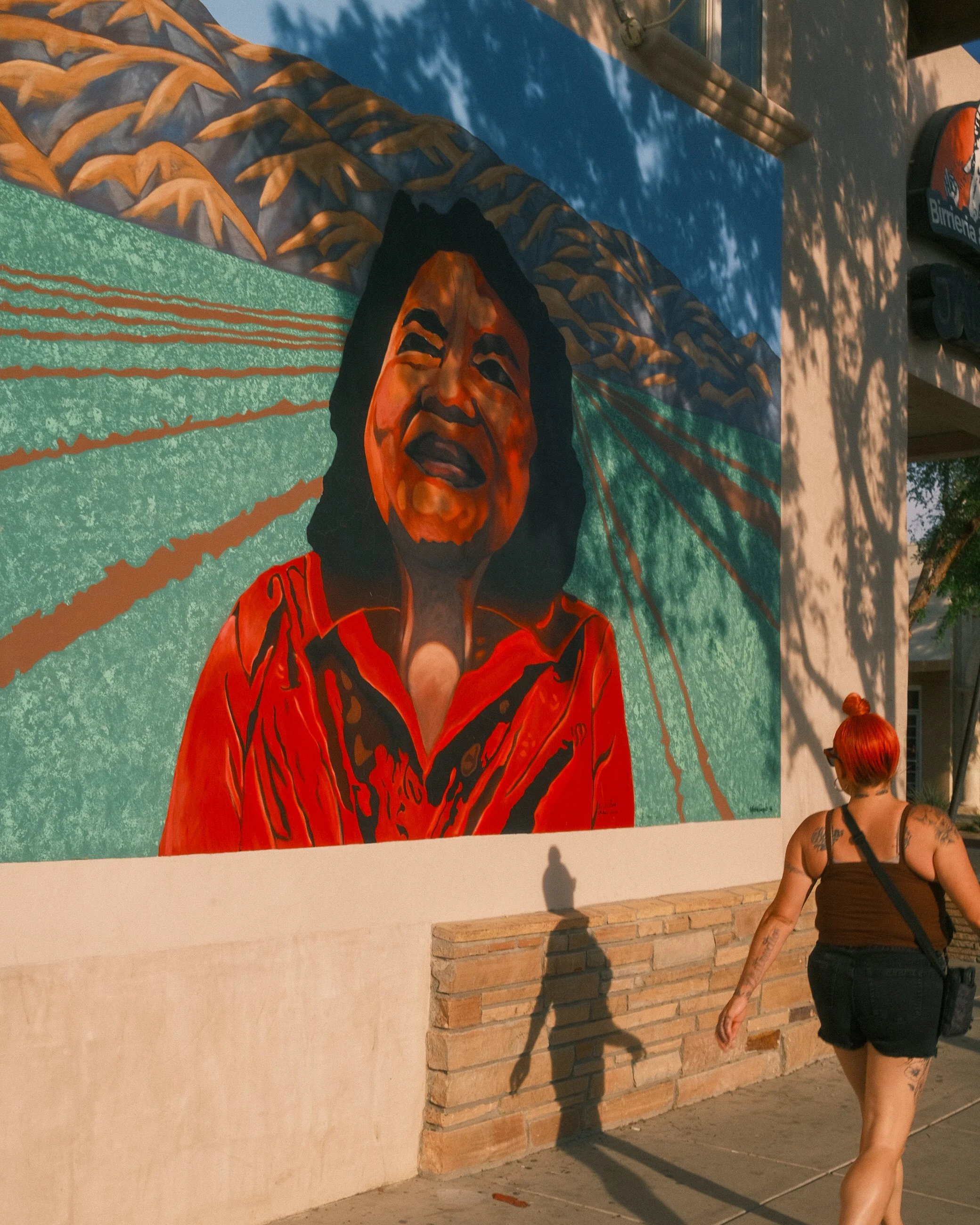 Large mural of a woman with open mouth, painted in vibrant colors, on an outdoor wall, with a person with red hair walking past casting a shadow.
