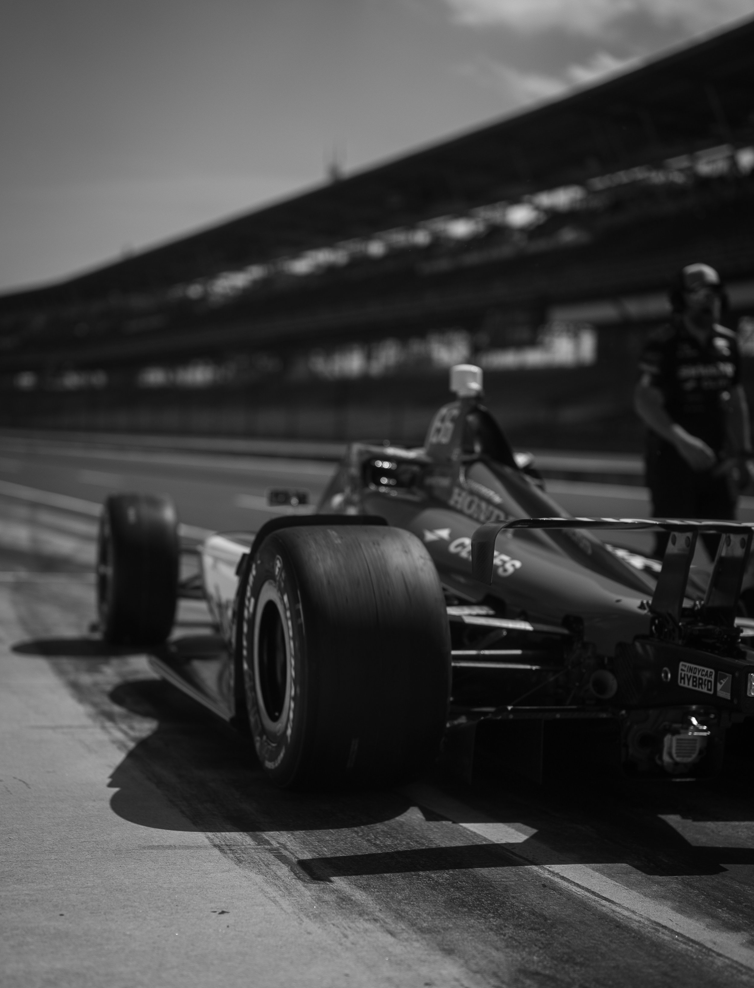 A black and white photograph of a Formula 1 race car on a racing track, with a person standing nearby and a grandstand in the background.