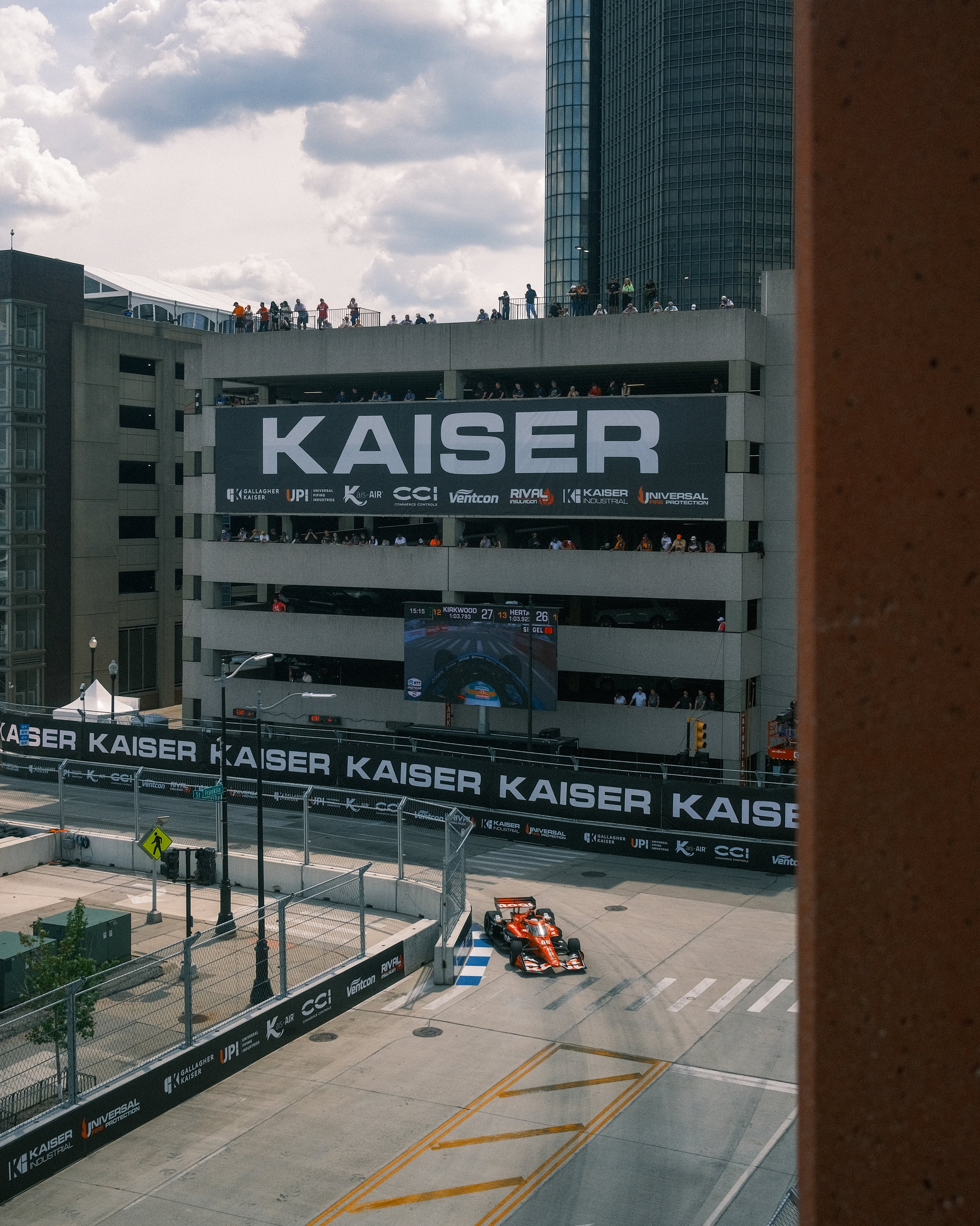 A racing car on a street circuit with a multi-level parking garage behind it displaying the word 'KAISER' and a large digital screen showing the race.