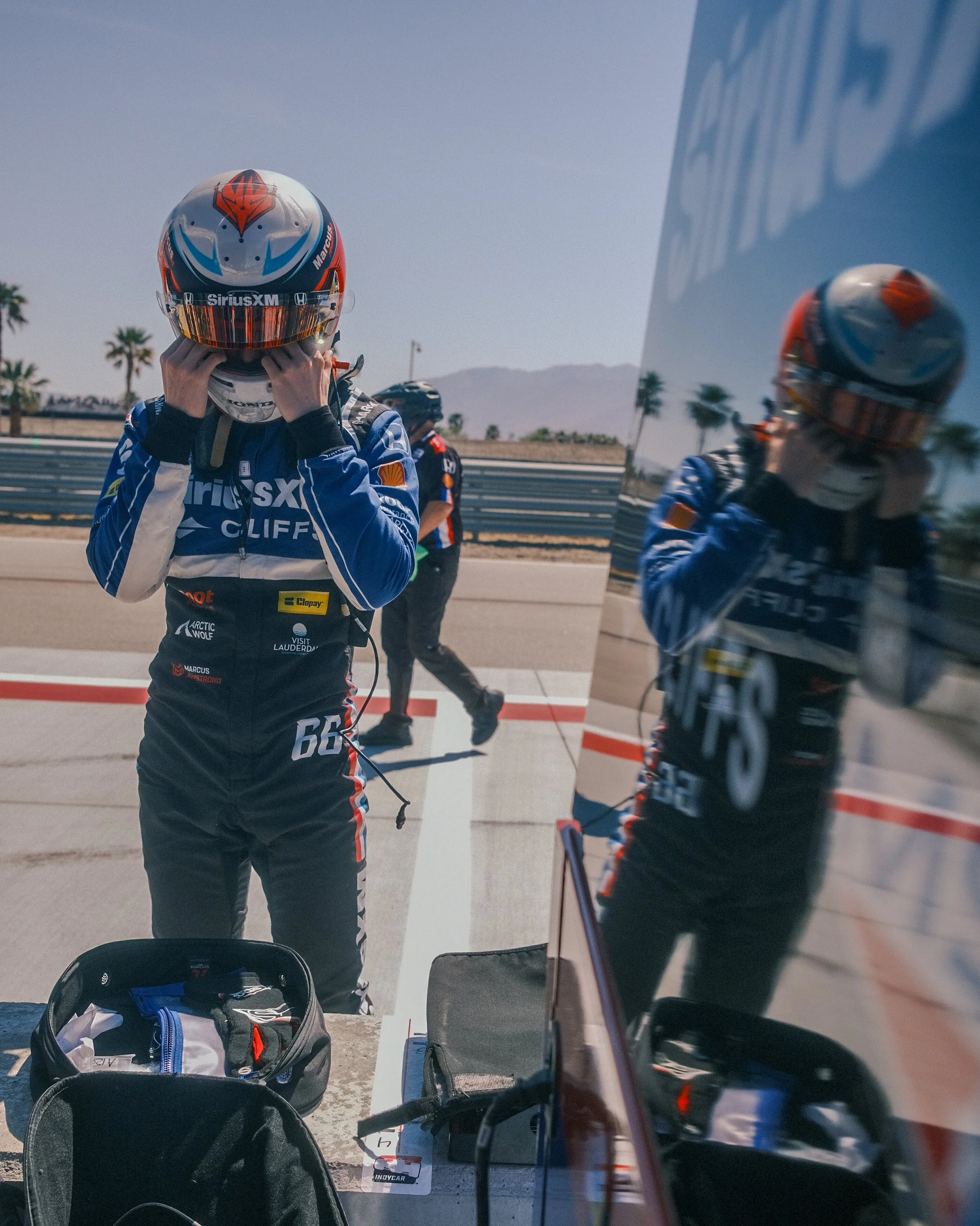 Race car driver in blue racing suit and helmet preparing in the pit area, with a reflective surface showing the driver and surroundings, desert landscape and mountains in the background.