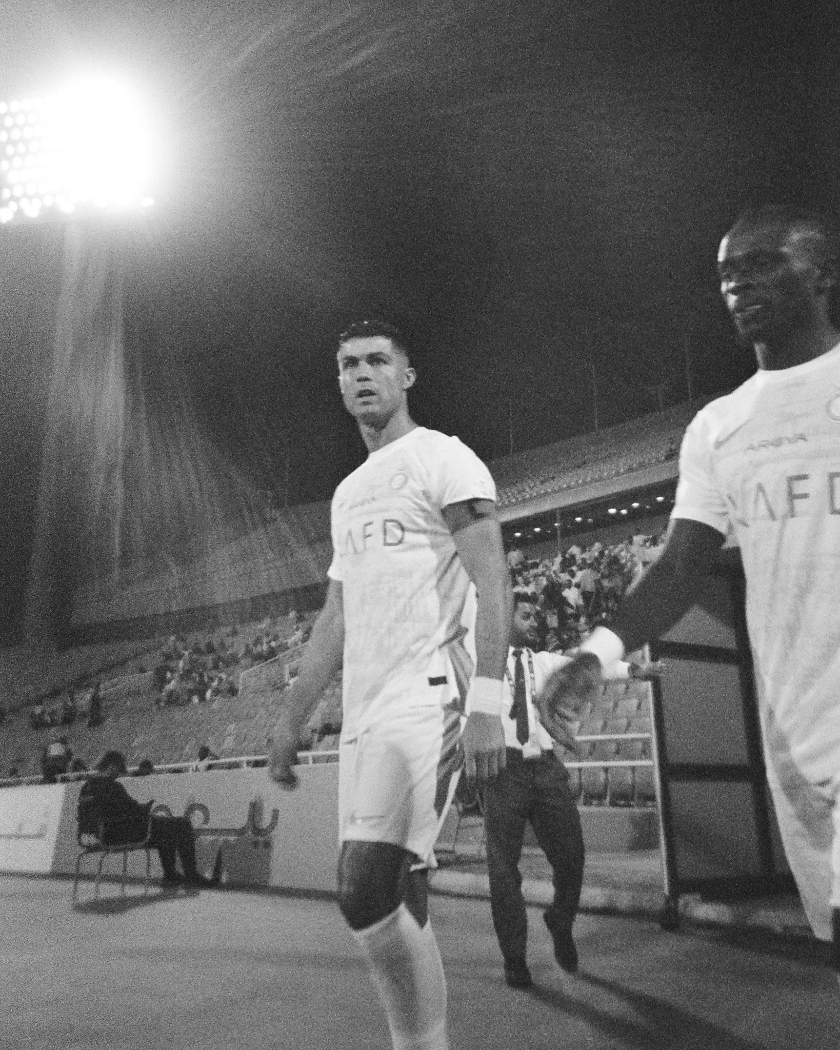 Two soccer players walking on the field during a game, with stadium seating and spectators in the background, black and white photo.