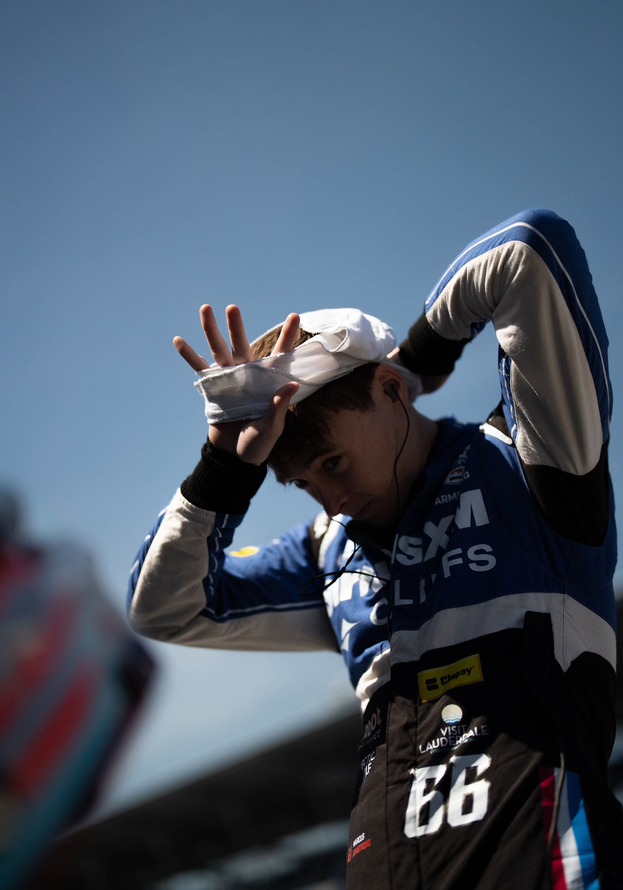 A young race car driver removes his helmet after a race, standing outdoors against a clear blue sky.