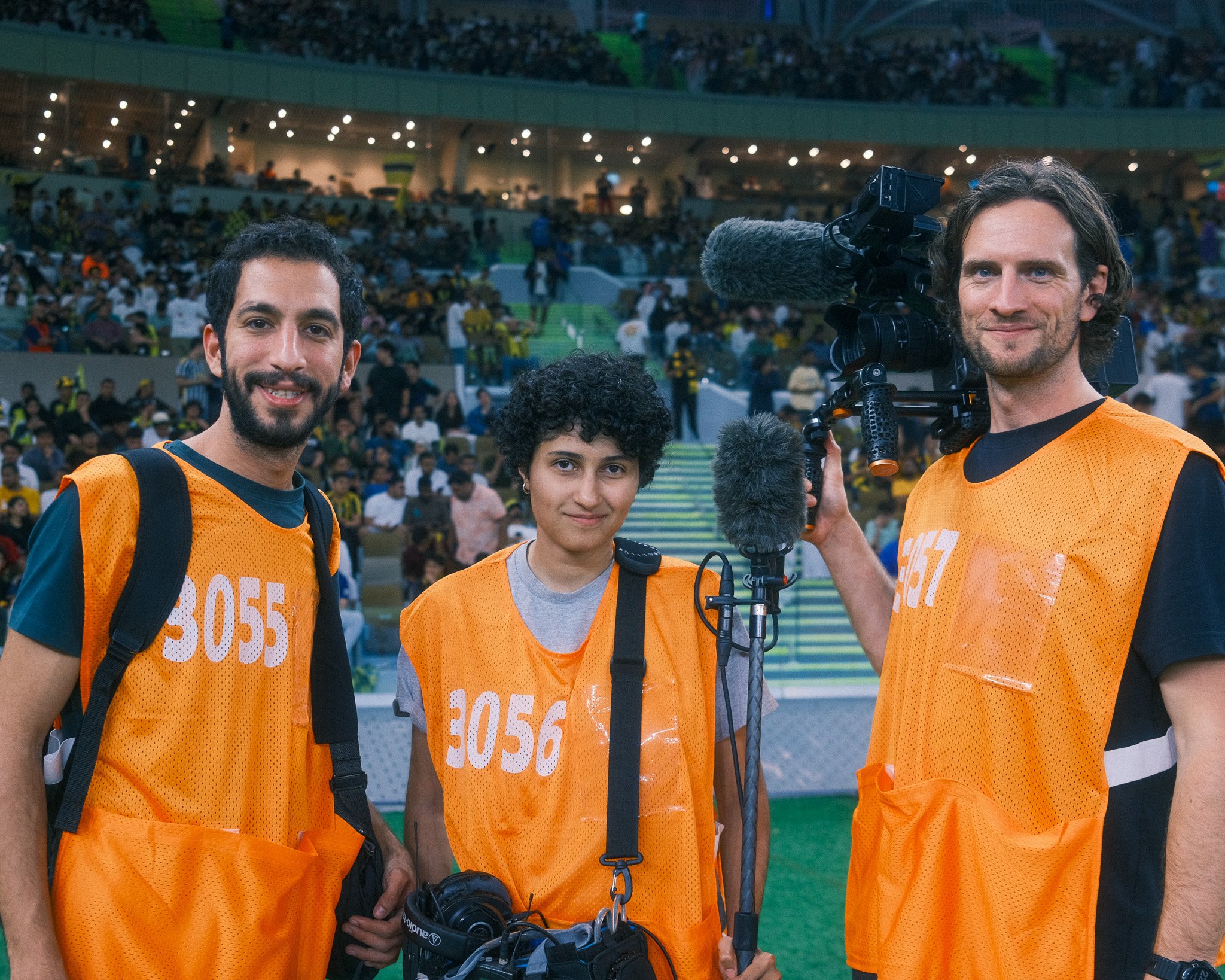 Three people wearing orange vests standing on a sports field with a crowd in the background. One person is holding a professional video camera, another has headphones, and the third has a backpack.