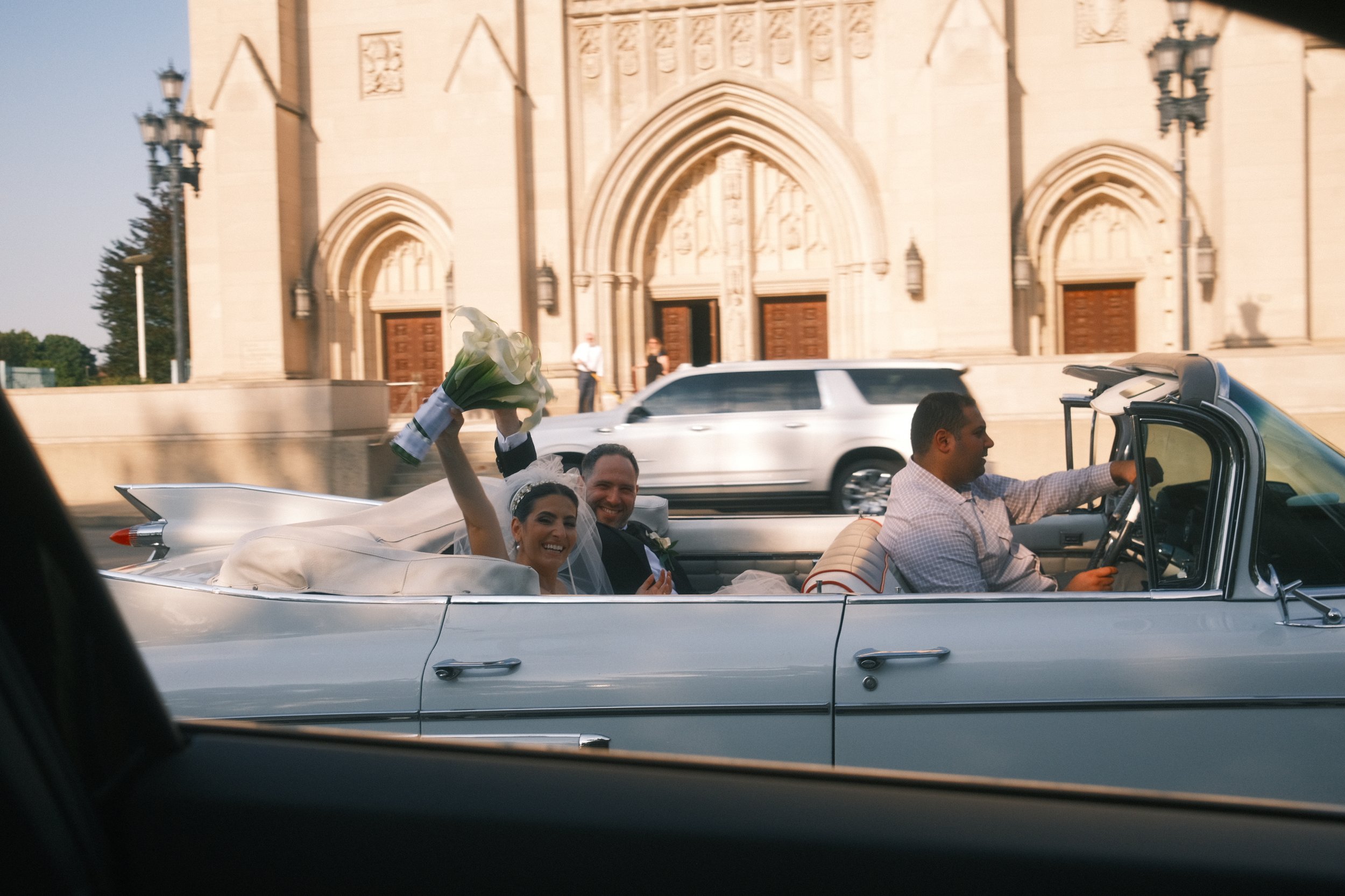 A bride and groom riding in a vintage convertible car, with the bride holding a bouquet and waving, in front of a church.