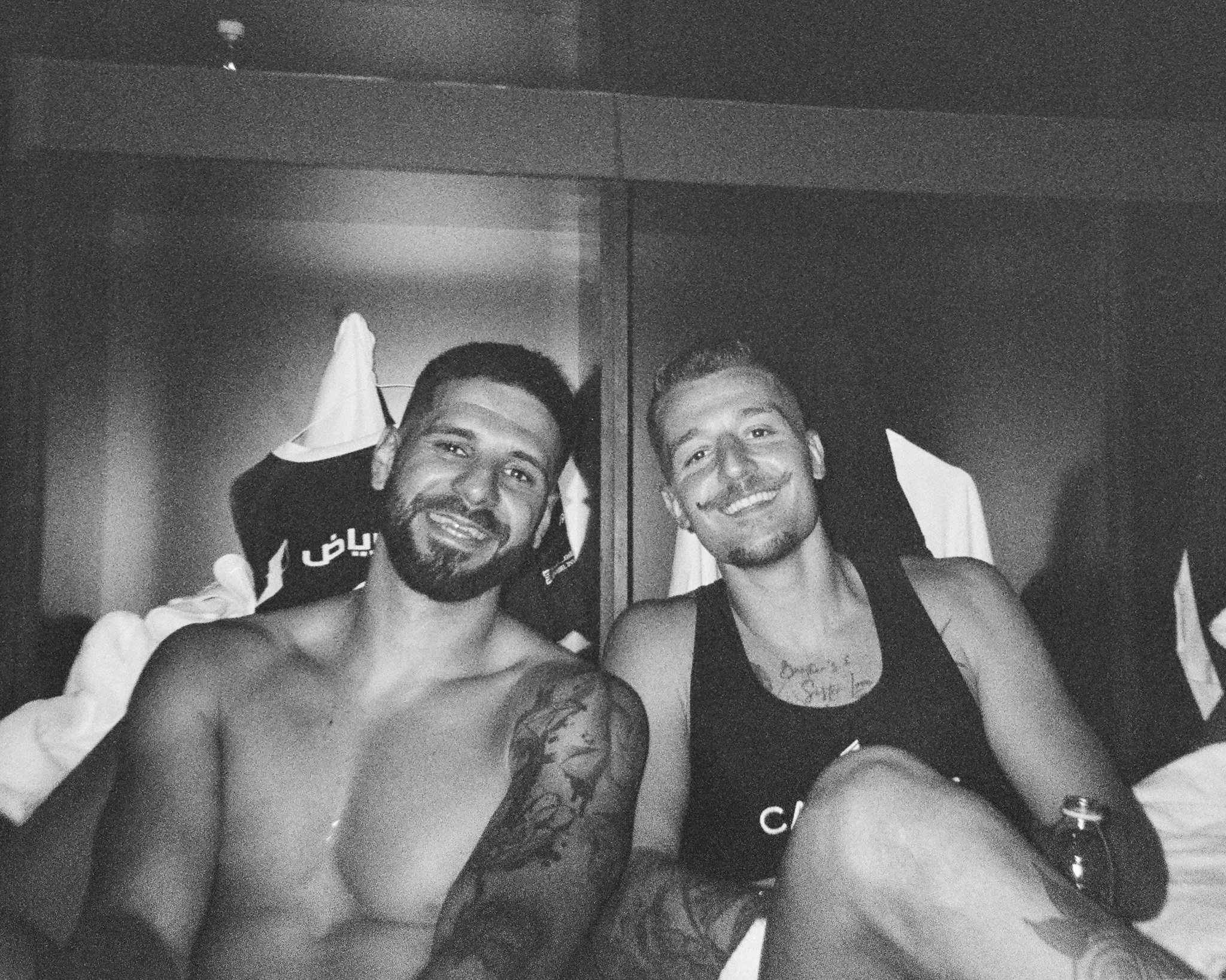 Two shirtless men smiling and sitting on a bed with pillows behind them in a dimly lit room.