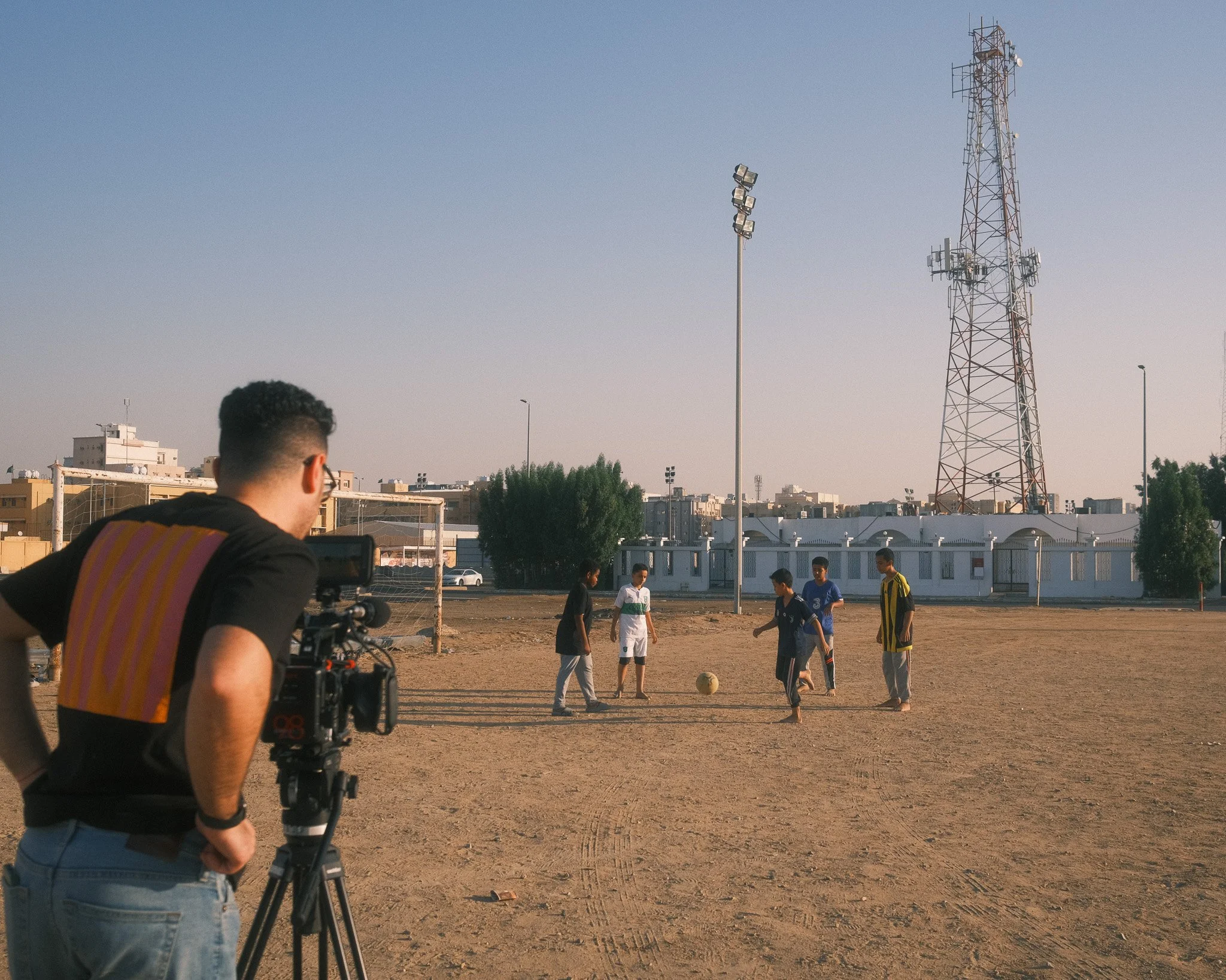 A man filming children playing soccer on an outdoor dirt field, with a camera on a tripod, in an urban setting during the daytime.