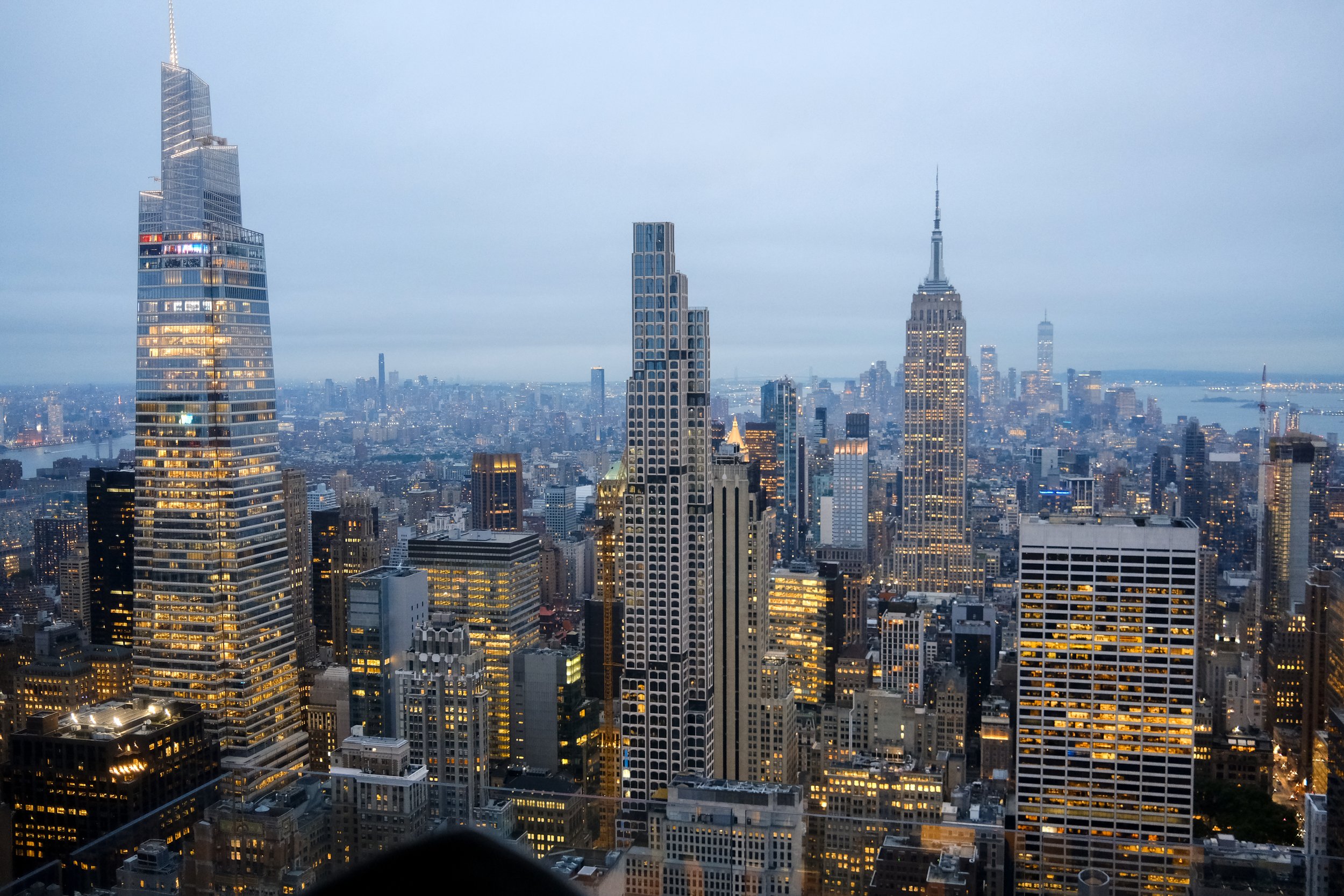 A city skyline of New York City with tall skyscrapers, including the Empire State Building, at dusk with illuminated windows.