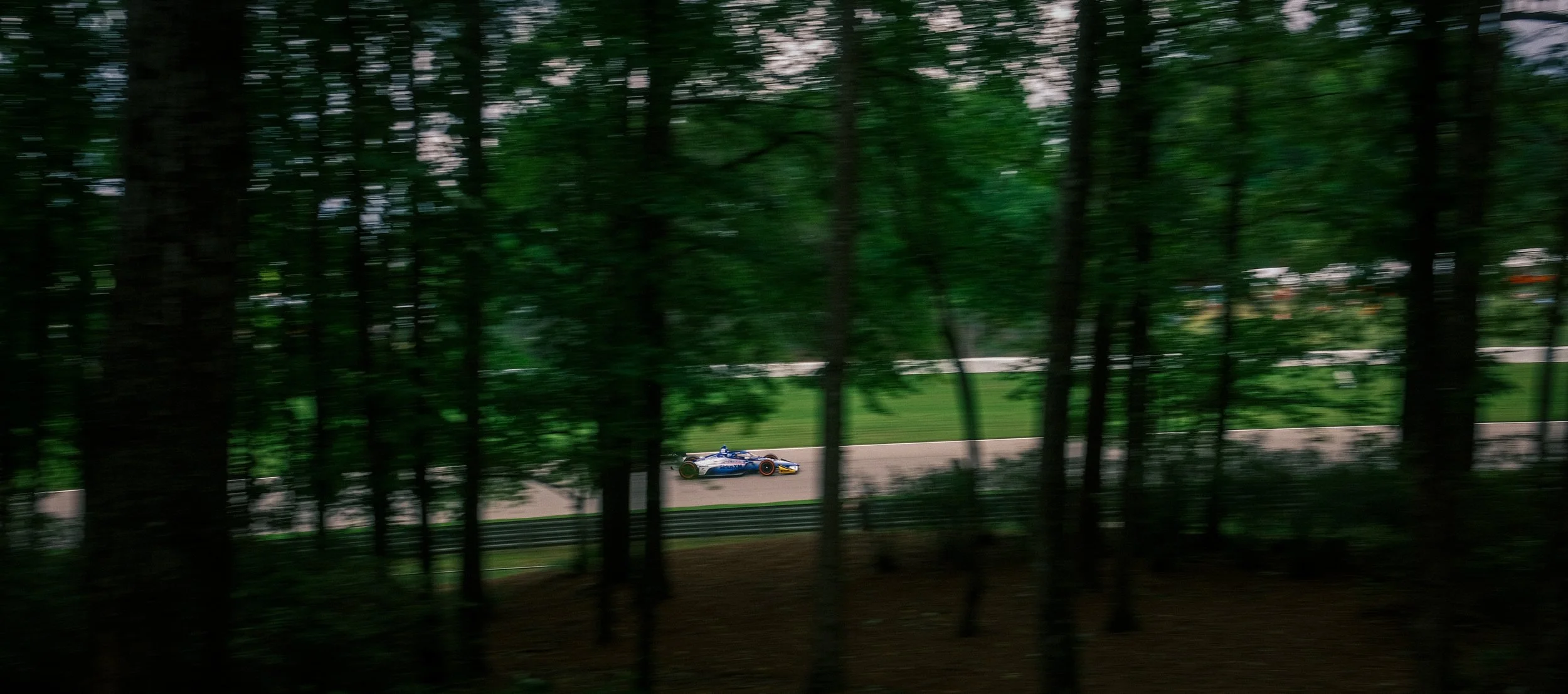 A race car on a track seen through a forest with trees and blurred greenery.