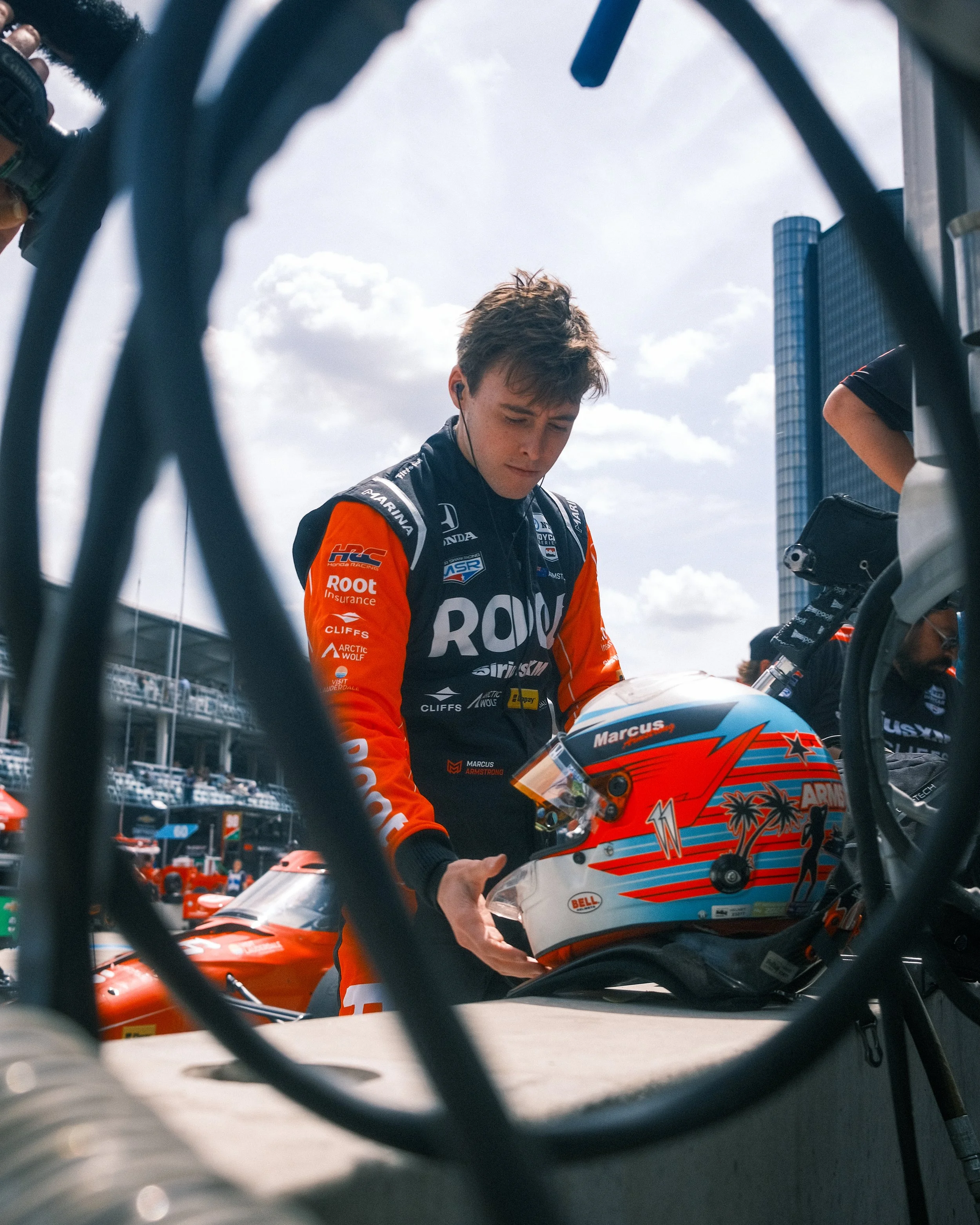 Race car driver in racing suit holding a helmet in a pit lane, with a race track and tall buildings in the background.