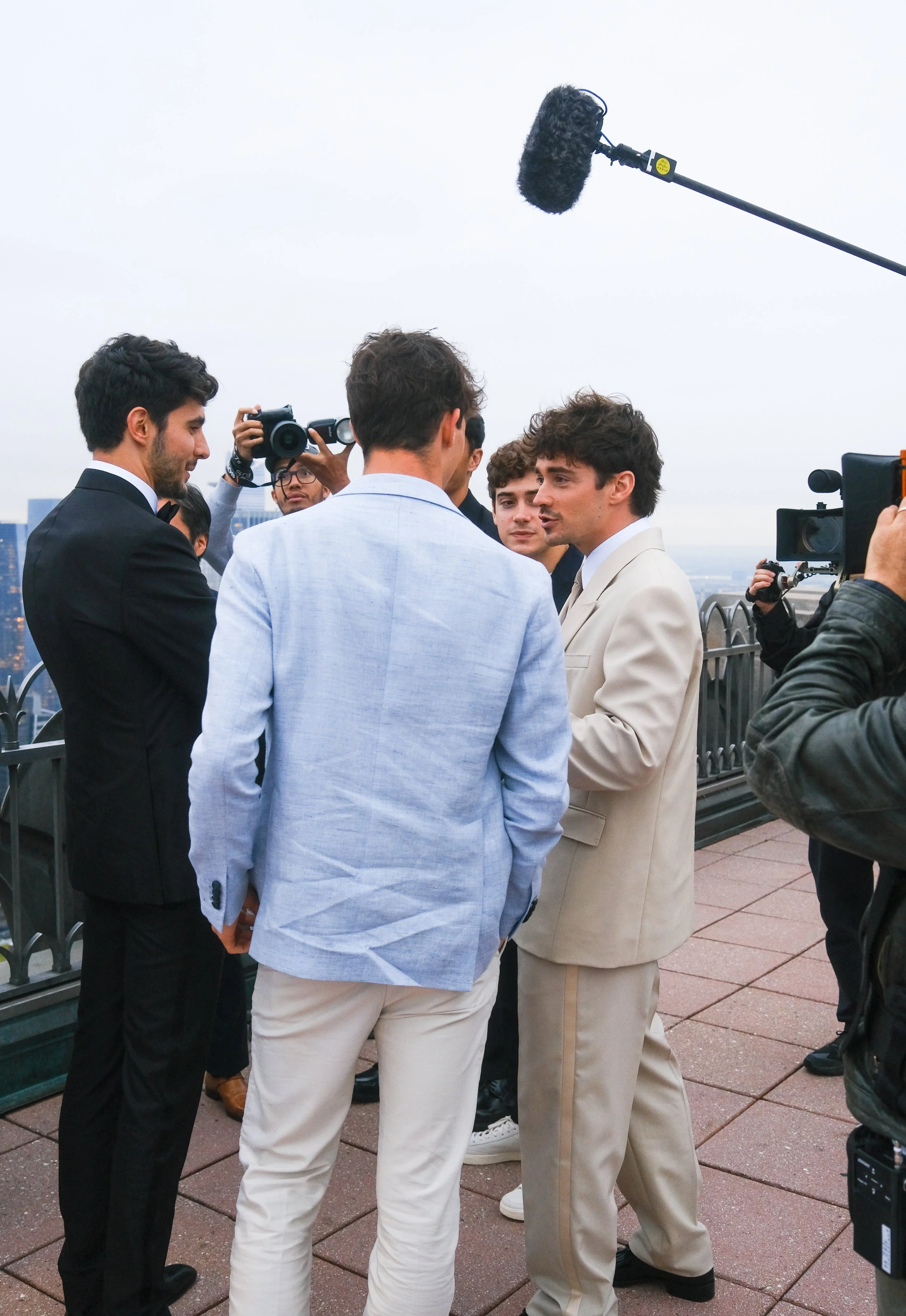 Group of men in suits being interviewed or photographed on a rooftop with city views, with professional camera operators and a microphone overhead.