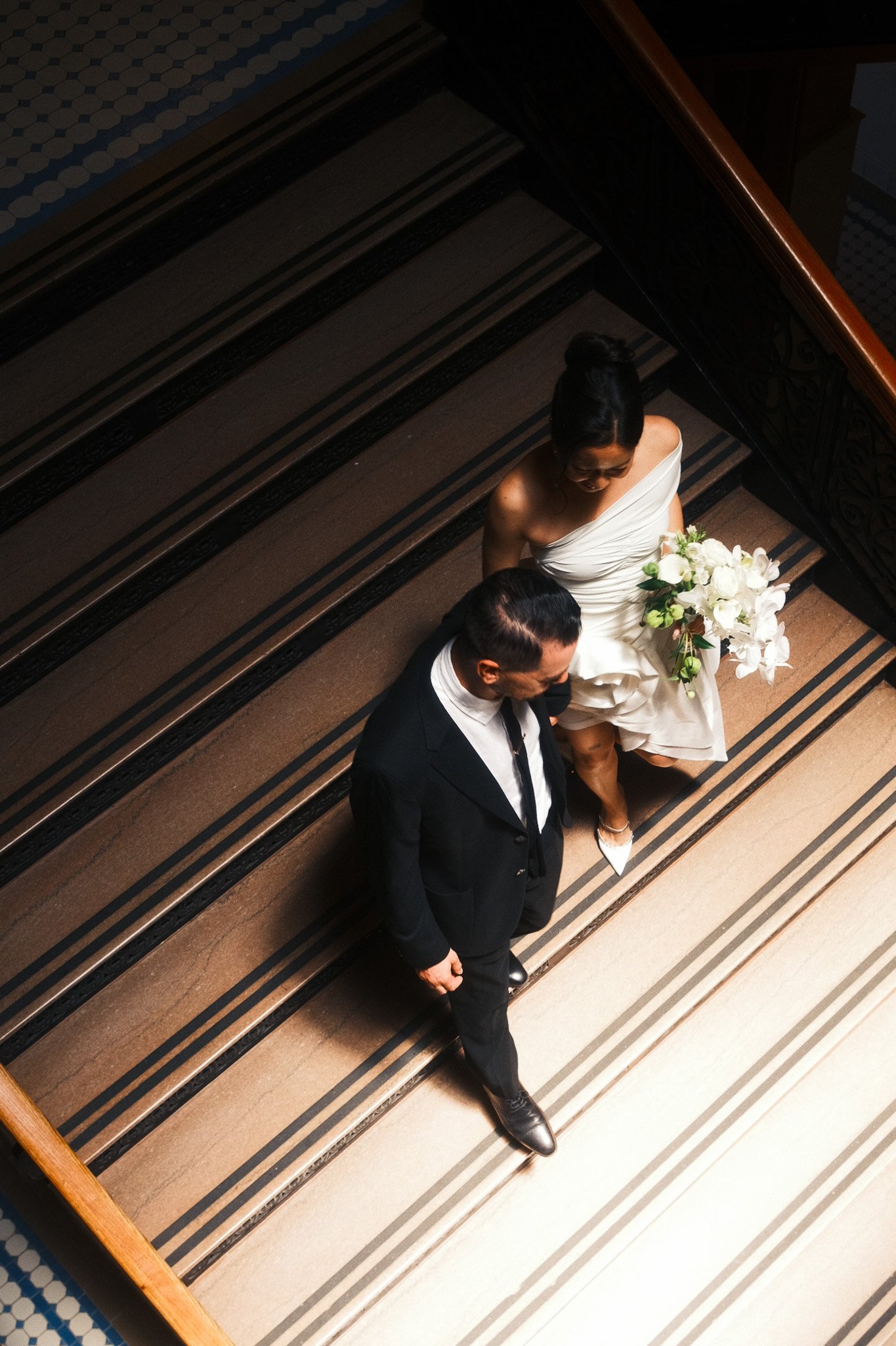 A couple in formal attire walking up a staircase. The woman is holding a bouquet of white flowers and wearing a white dress. The man is dressed in a black suit with a white shirt and tie.