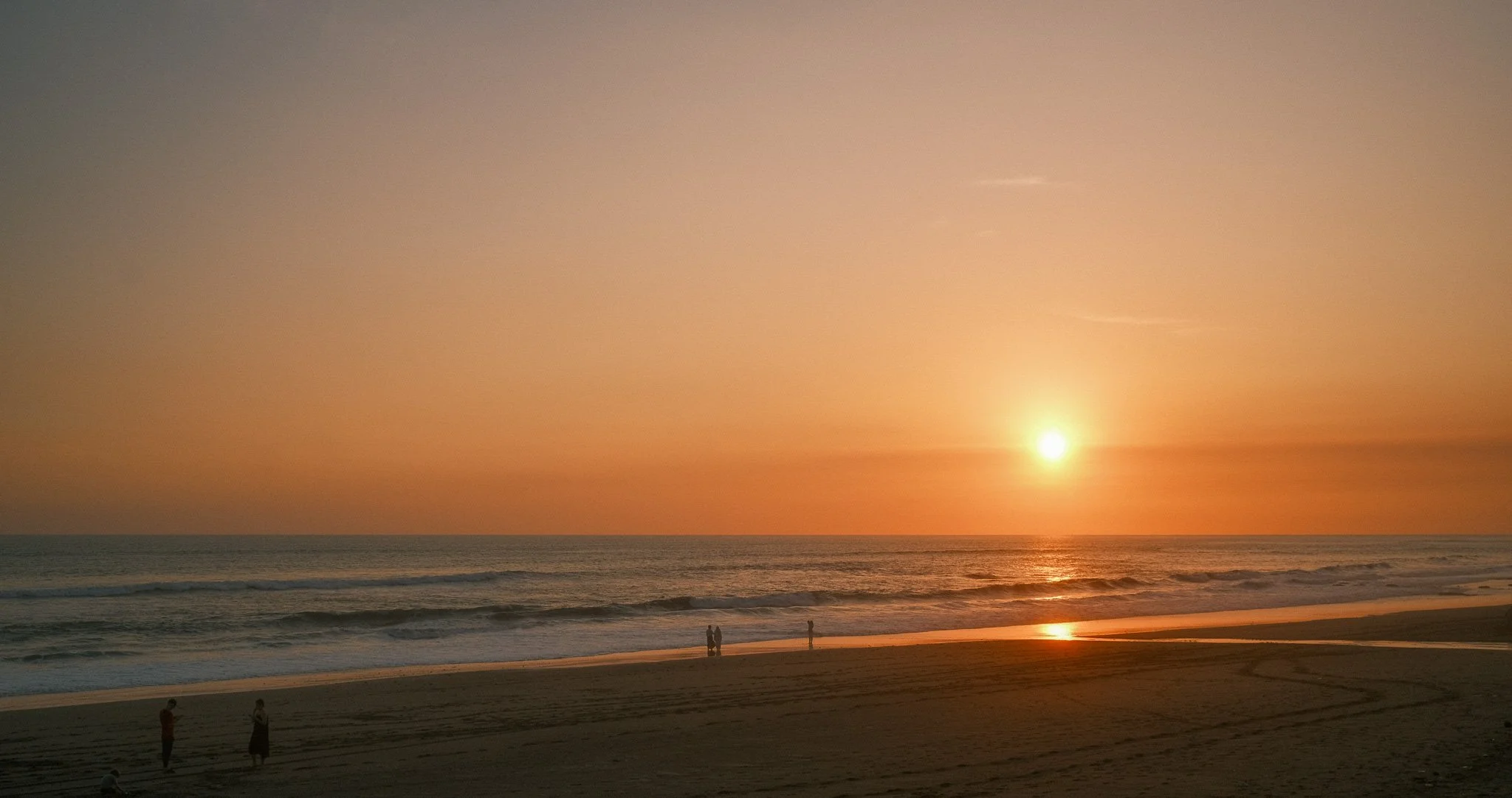 A beach at sunset with the sun near the horizon, casting a warm glow on the ocean and sand. Small groups of people are walking and standing along the shoreline, enjoying the peaceful scene.