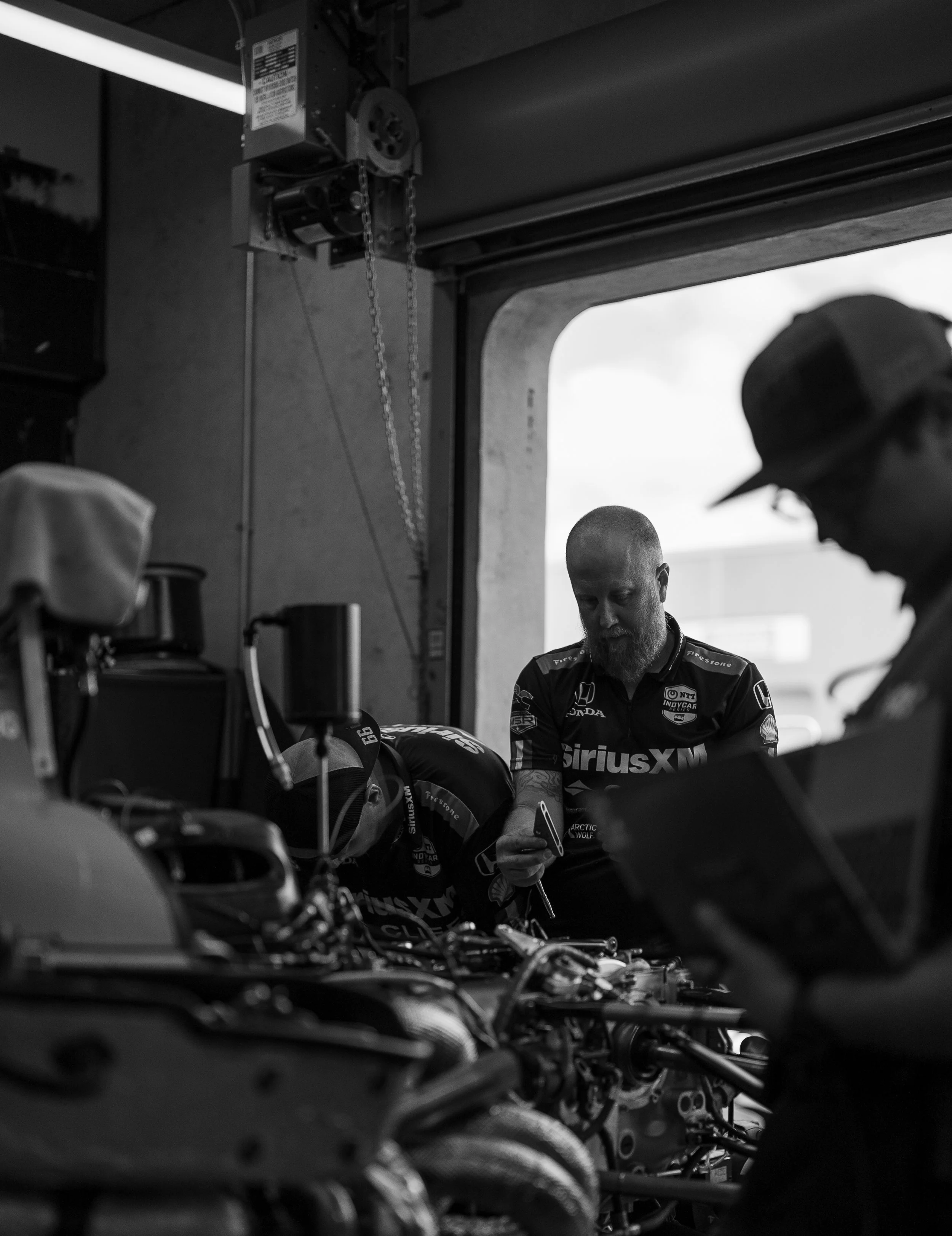 Black and white photo of two mechanics working on a race car inside a garage, one holding a tool, with racing team logos on their uniforms.