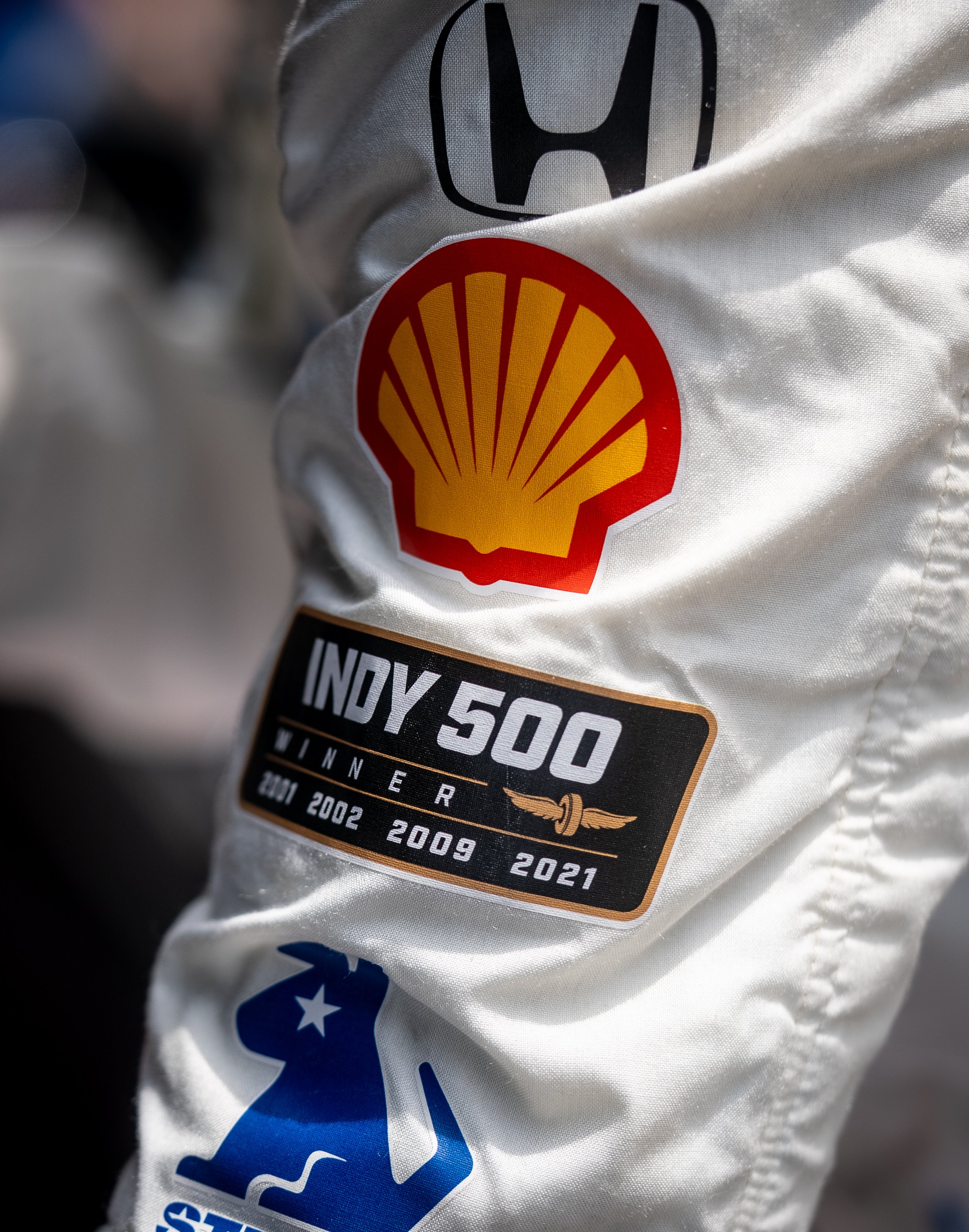 Close-up of a racing suit sleeve with logos including Honda, Shell, Indy 500 winner from 2001 to 2021, and a blue logo with a stylized dog.