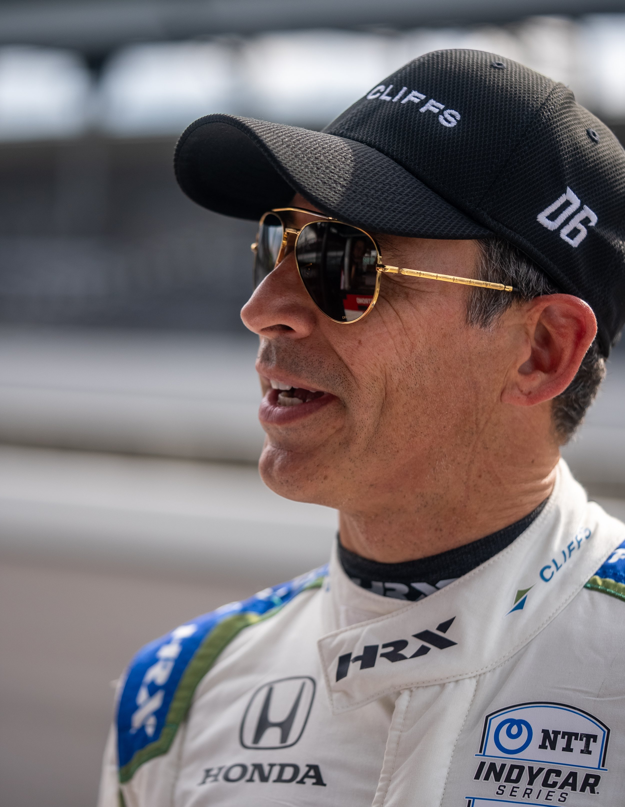 A man wearing a black cap with 'CLIFRS' and '06' embroidered on it, sunglasses, and a racing suit with various sponsor logos, including Honda and NTT IndyCar Series.