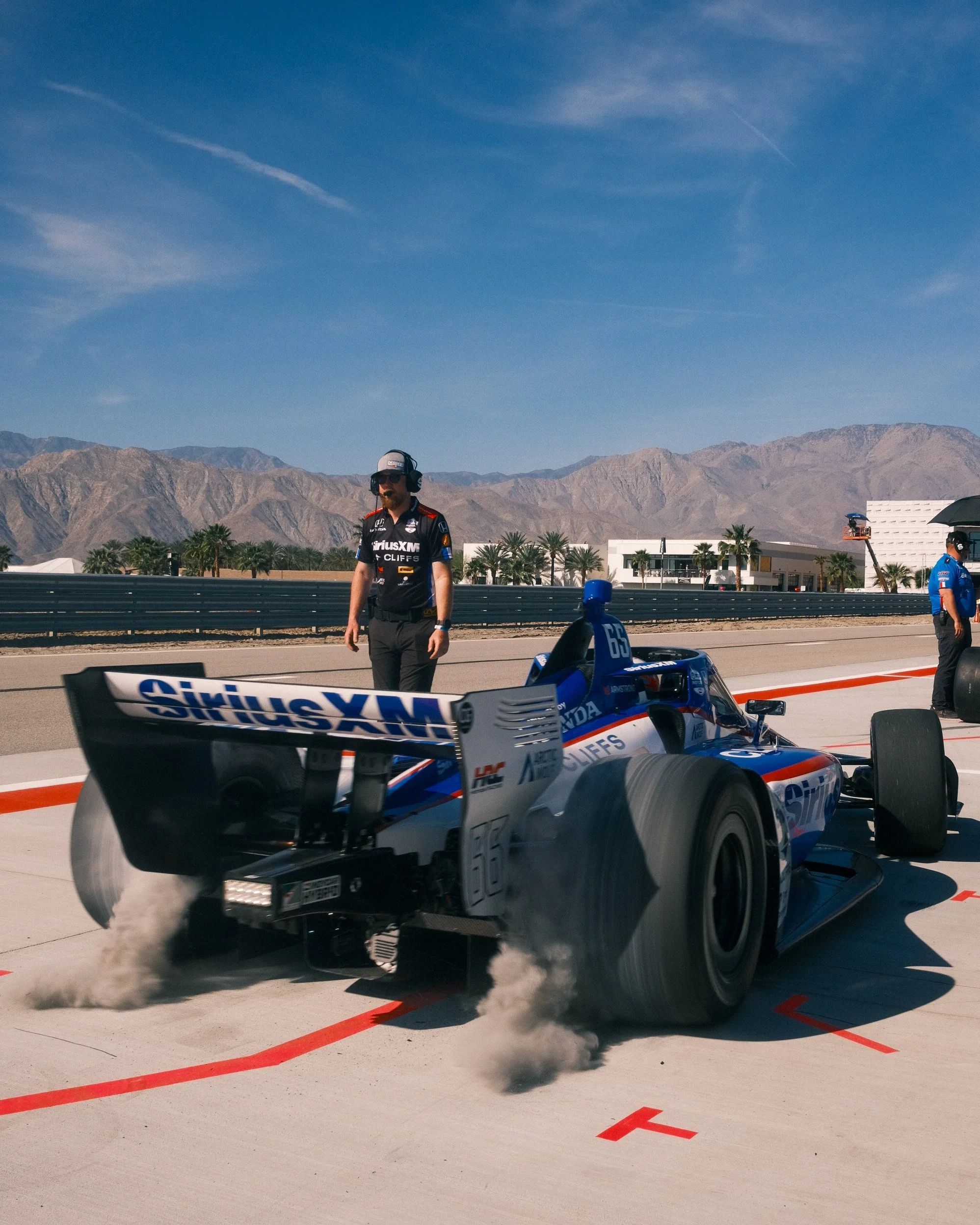 IndyCar race car in the pit lane with crew members, one standing and another holding an umbrella, in a desert setting with mountains in the background.