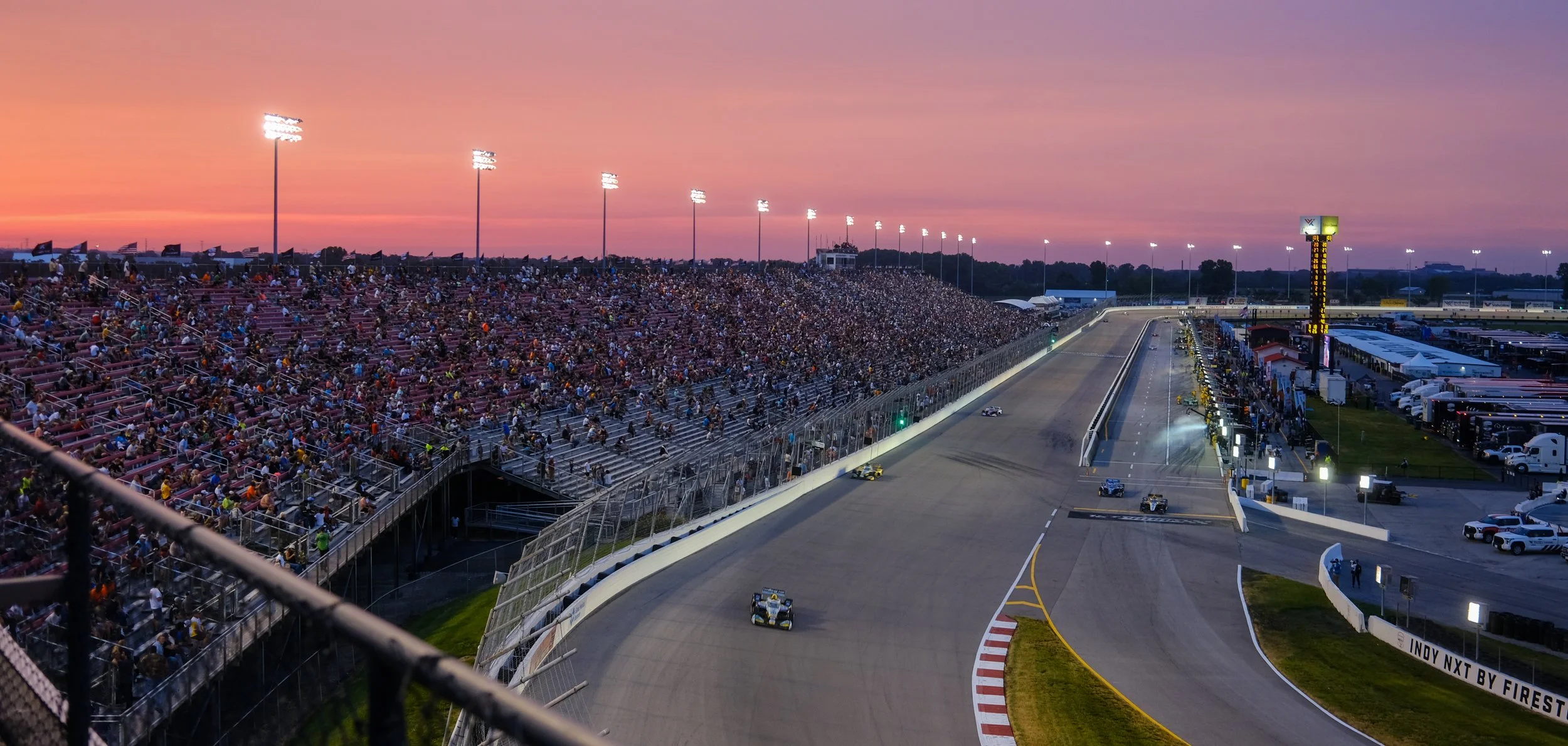 Sunset at a car racing track with a large crowd in the grandstand, race cars on the track, and bright stadium lights illuminating the scene.