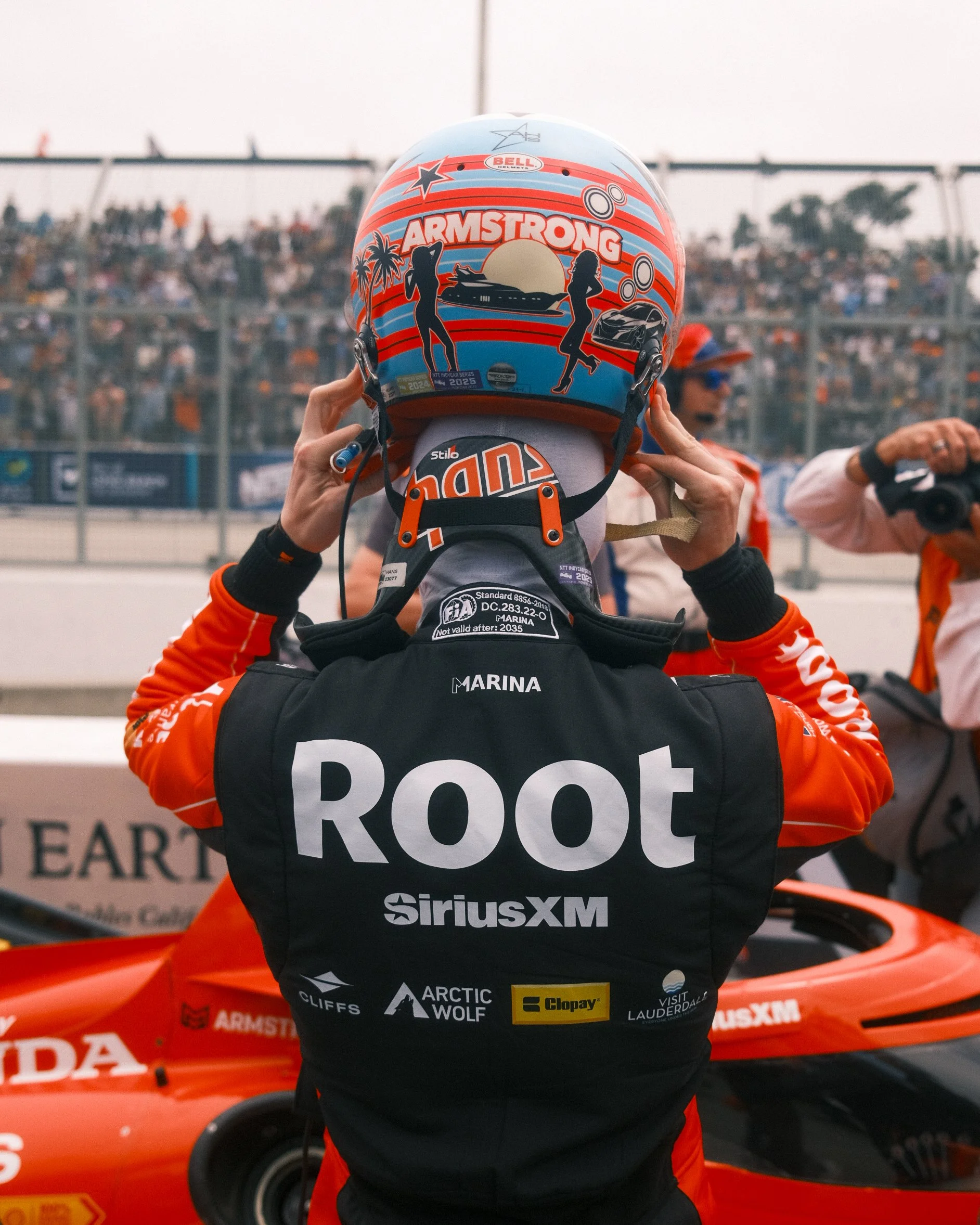 Race car driver adjusting helmet with a colorful design, standing in front of a race car at a racetrack, with spectators in the background.