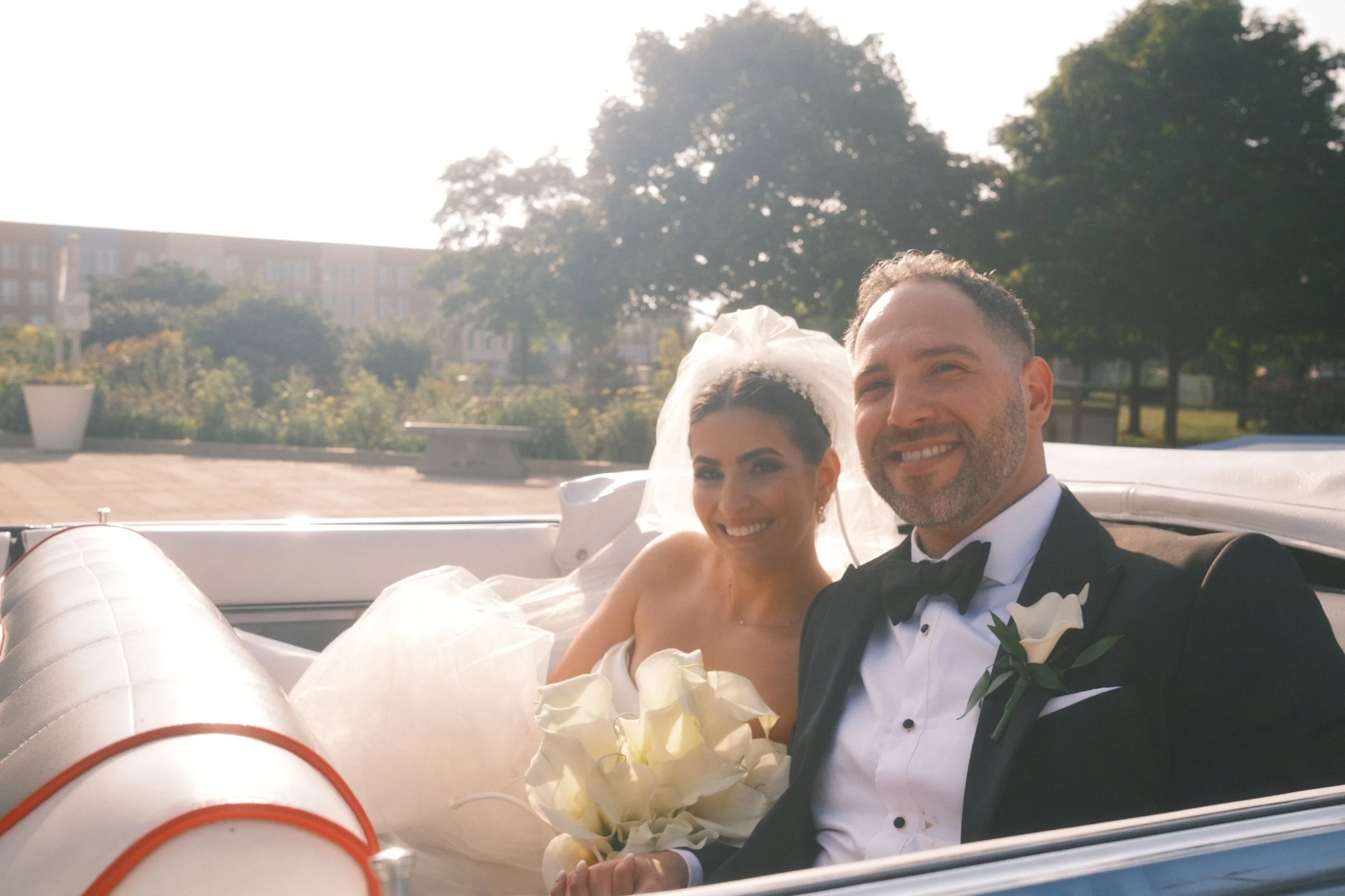A newlywed couple in wedding attire riding in a vintage car, smiling and holding a bouquet of white calla lilies, outdoors on a sunny day with trees and buildings in the background.