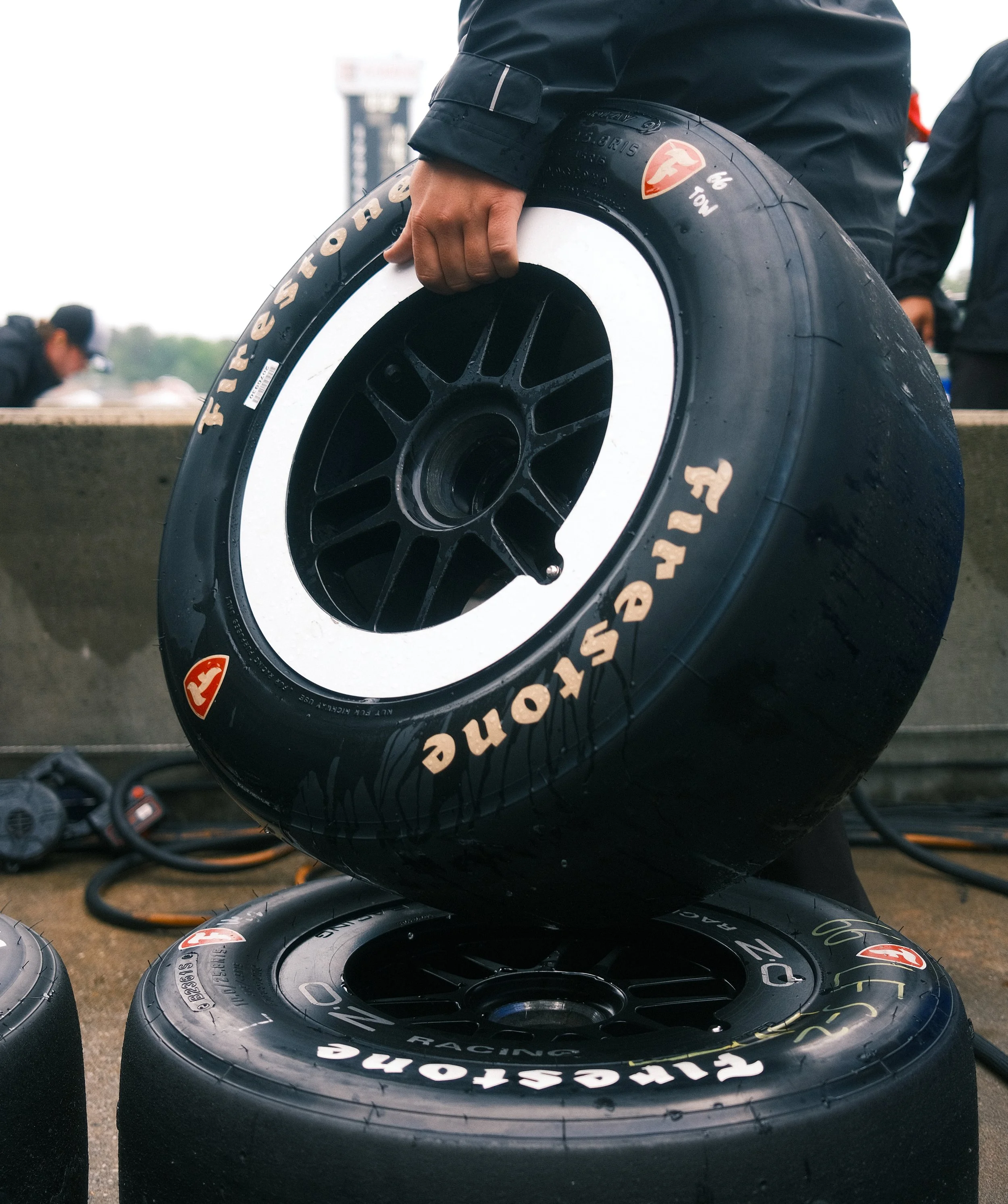 Person holding racing tire with another tire on the ground, at a race track.