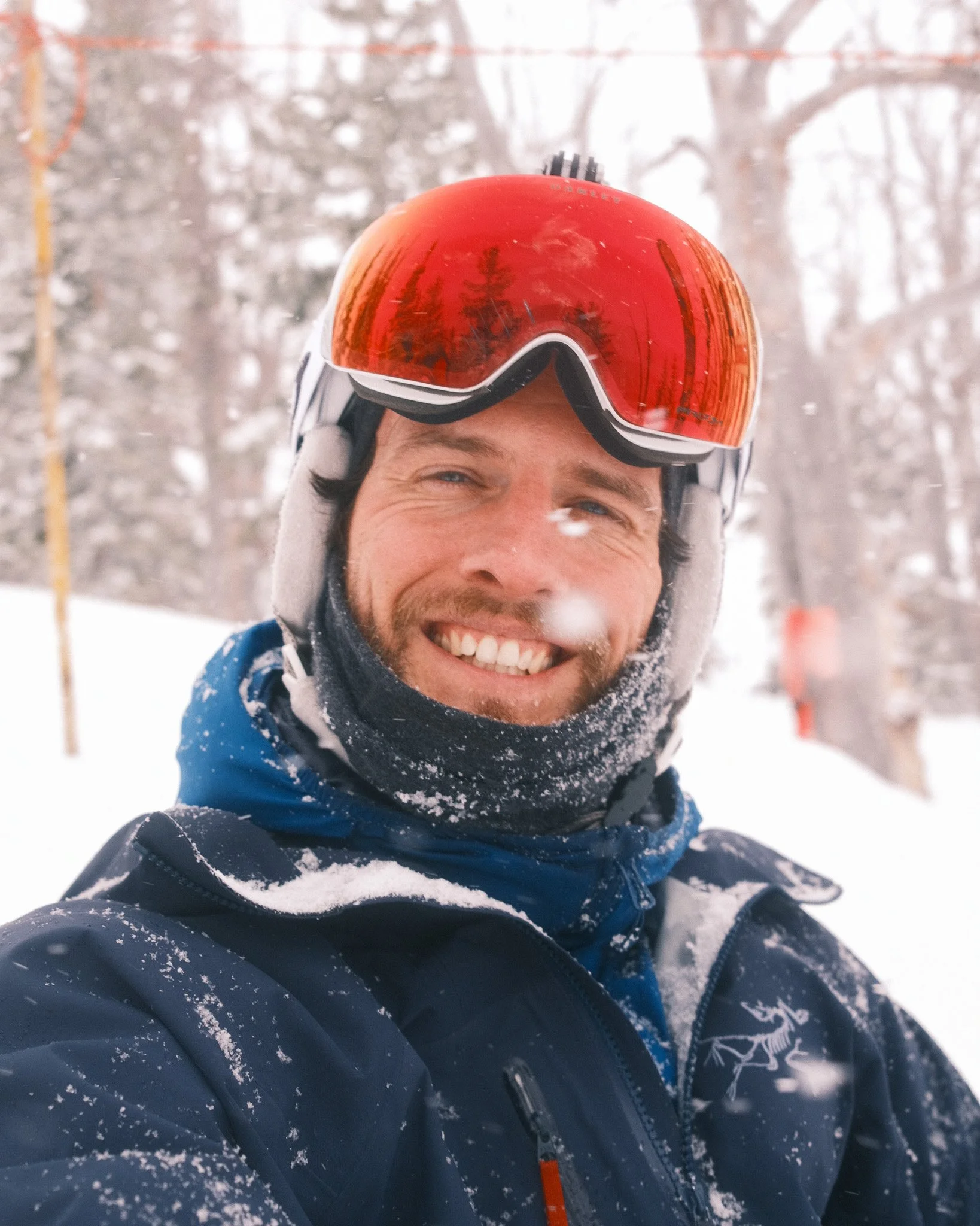 A man in snow gear takes a selfie in snowy woods, wearing a red ski helmet and goggles, smiling with snow on his jacket and face.