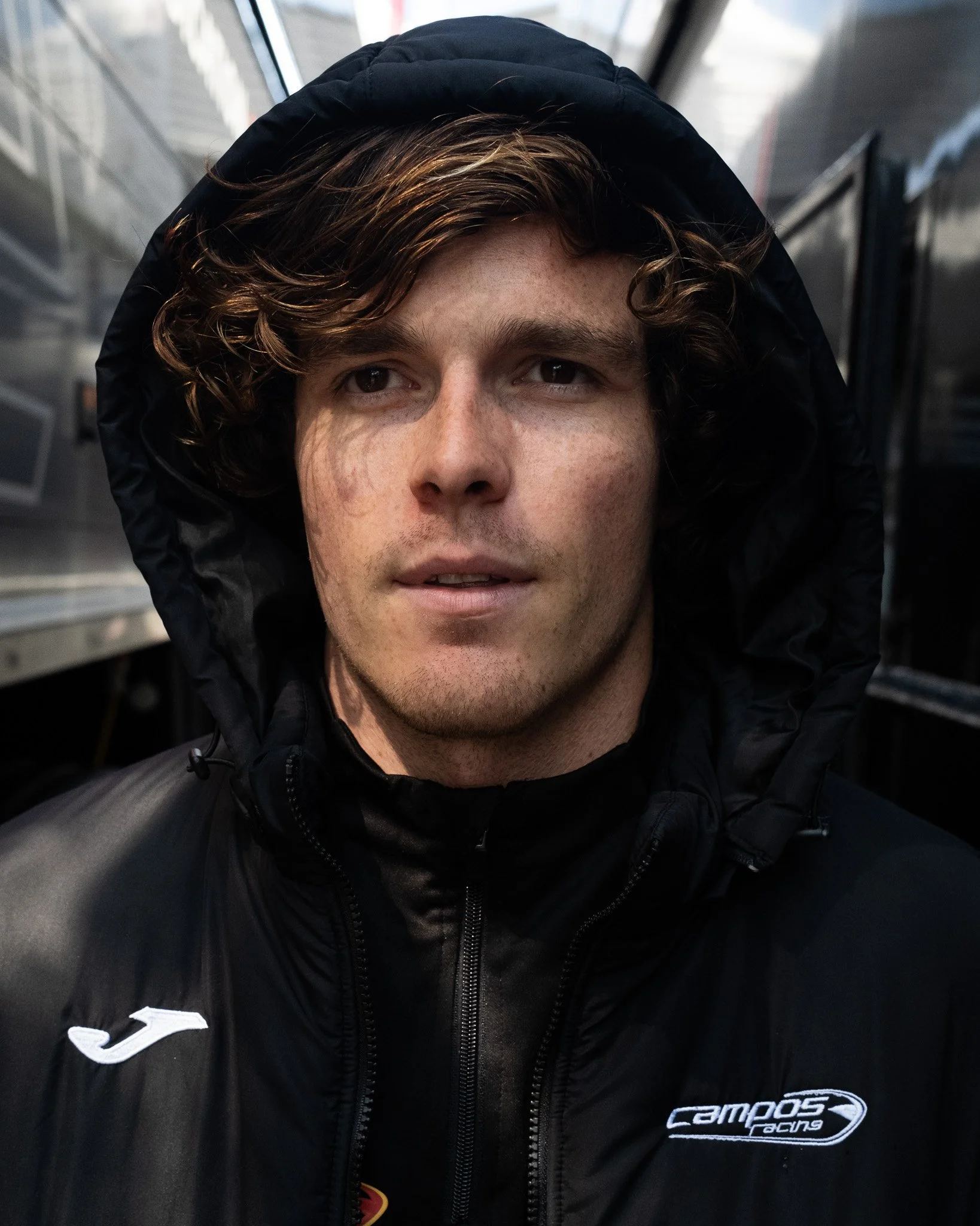 A young man with wavy brown hair wearing a black hooded jacket in a close-up portrait.