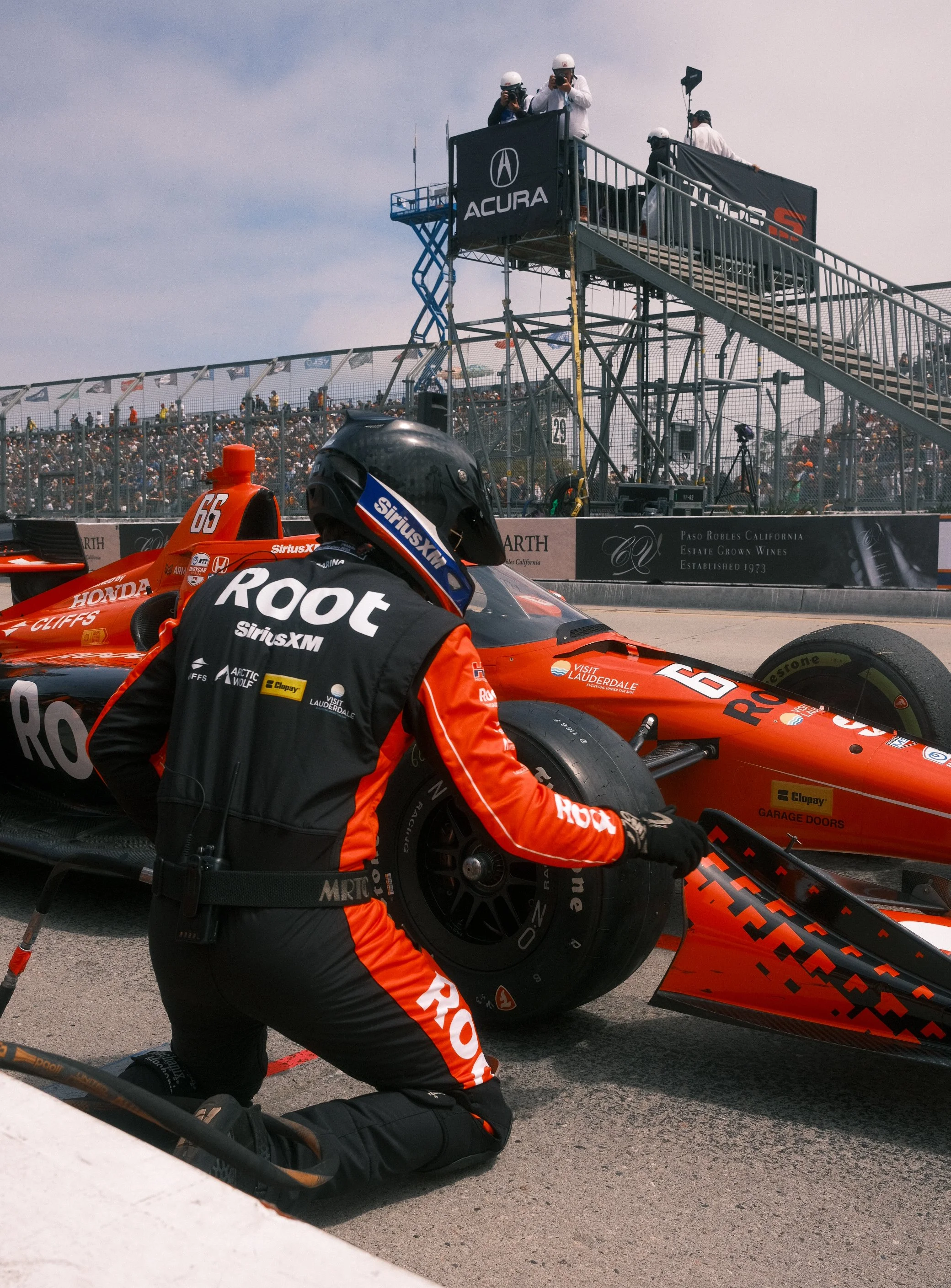 A race car driver kneeling next to a red IndyCar during a pit stop at a racetrack, with crew members in the background on a pit wall and spectators in the stands.