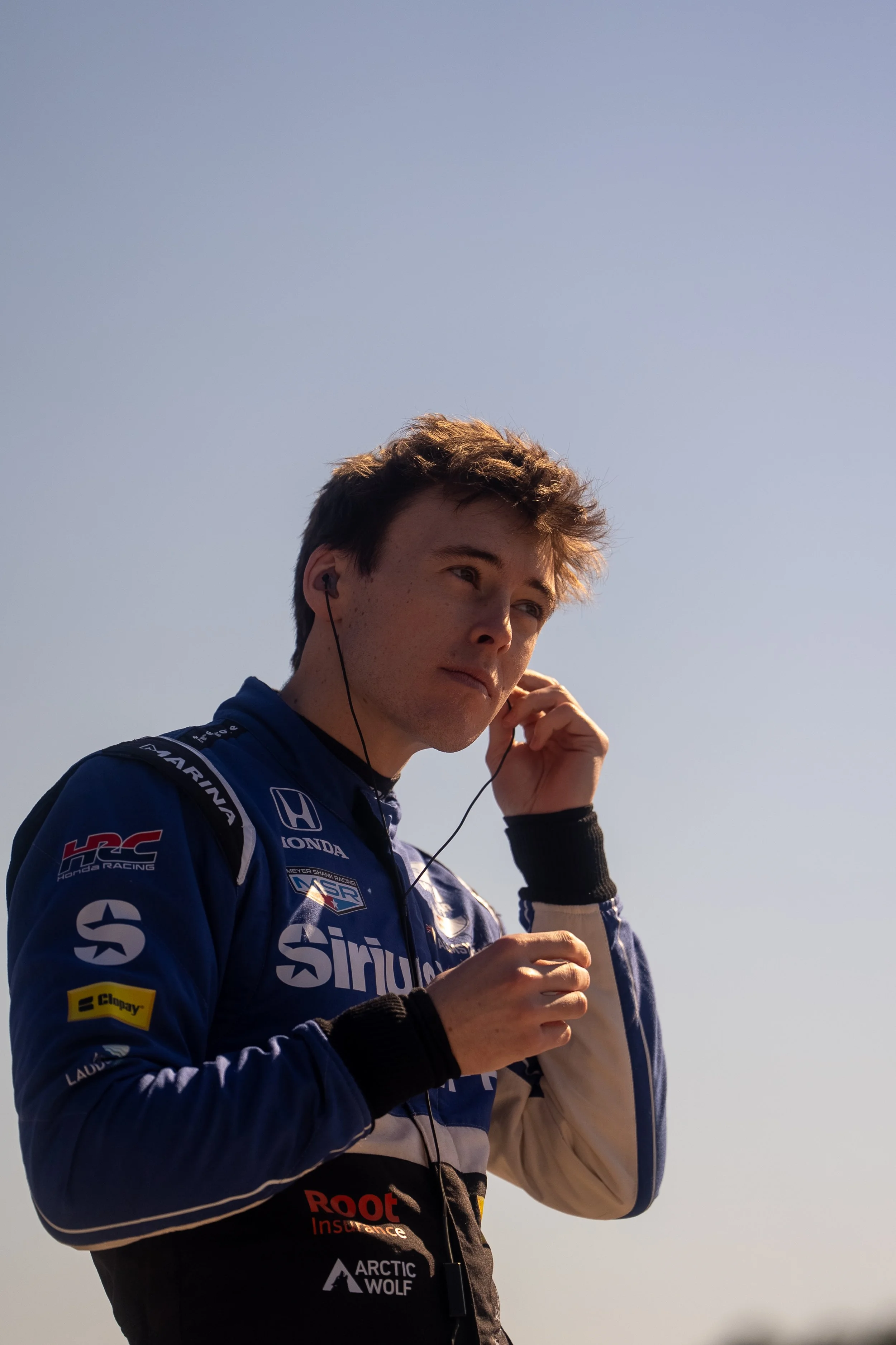 Young male race car driver in a blue racing suit with sponsor logos, adjusting his earphones against a clear sky.