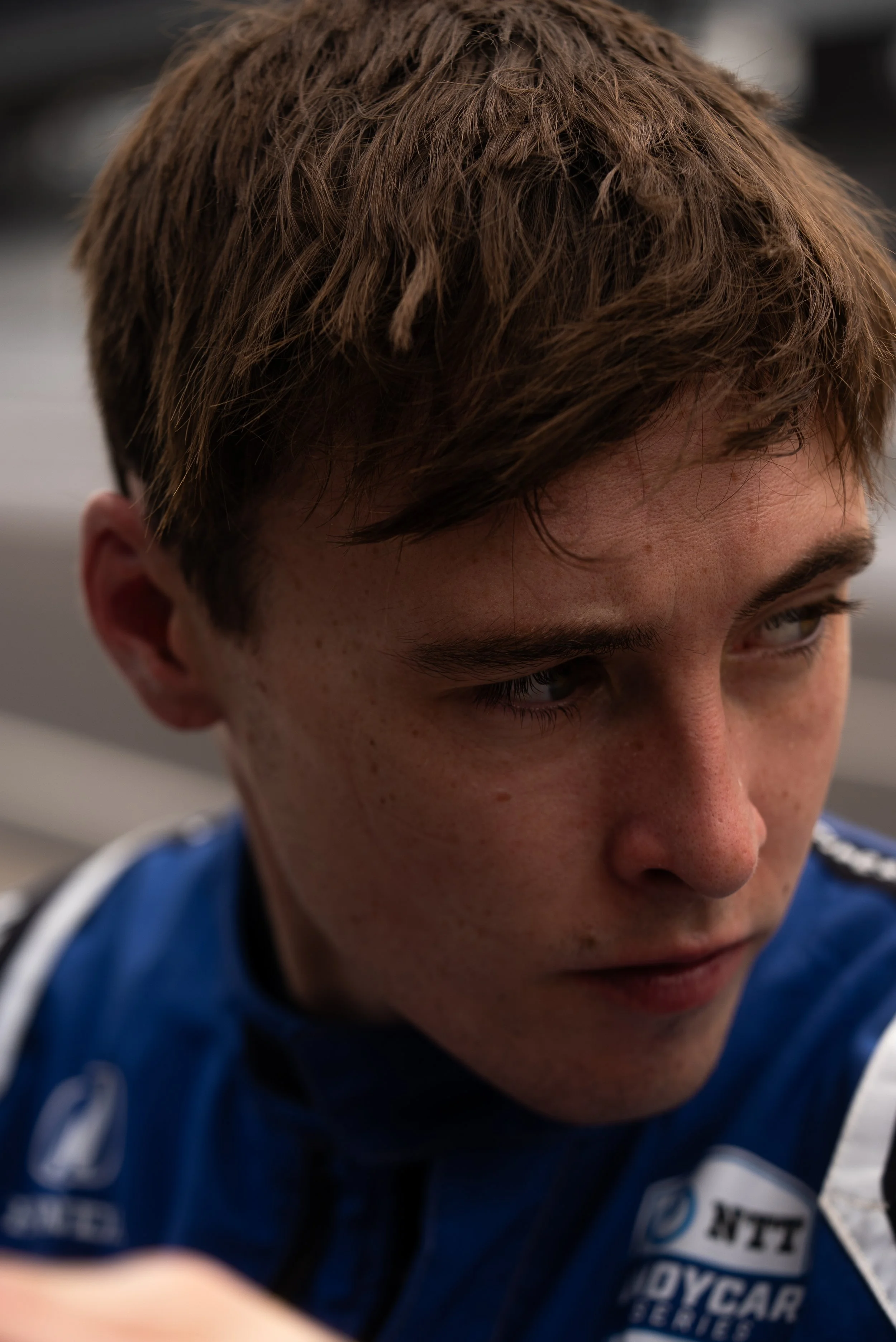 Close-up of a young man with brown hair and freckles, wearing a blue racing suit, looking downward.