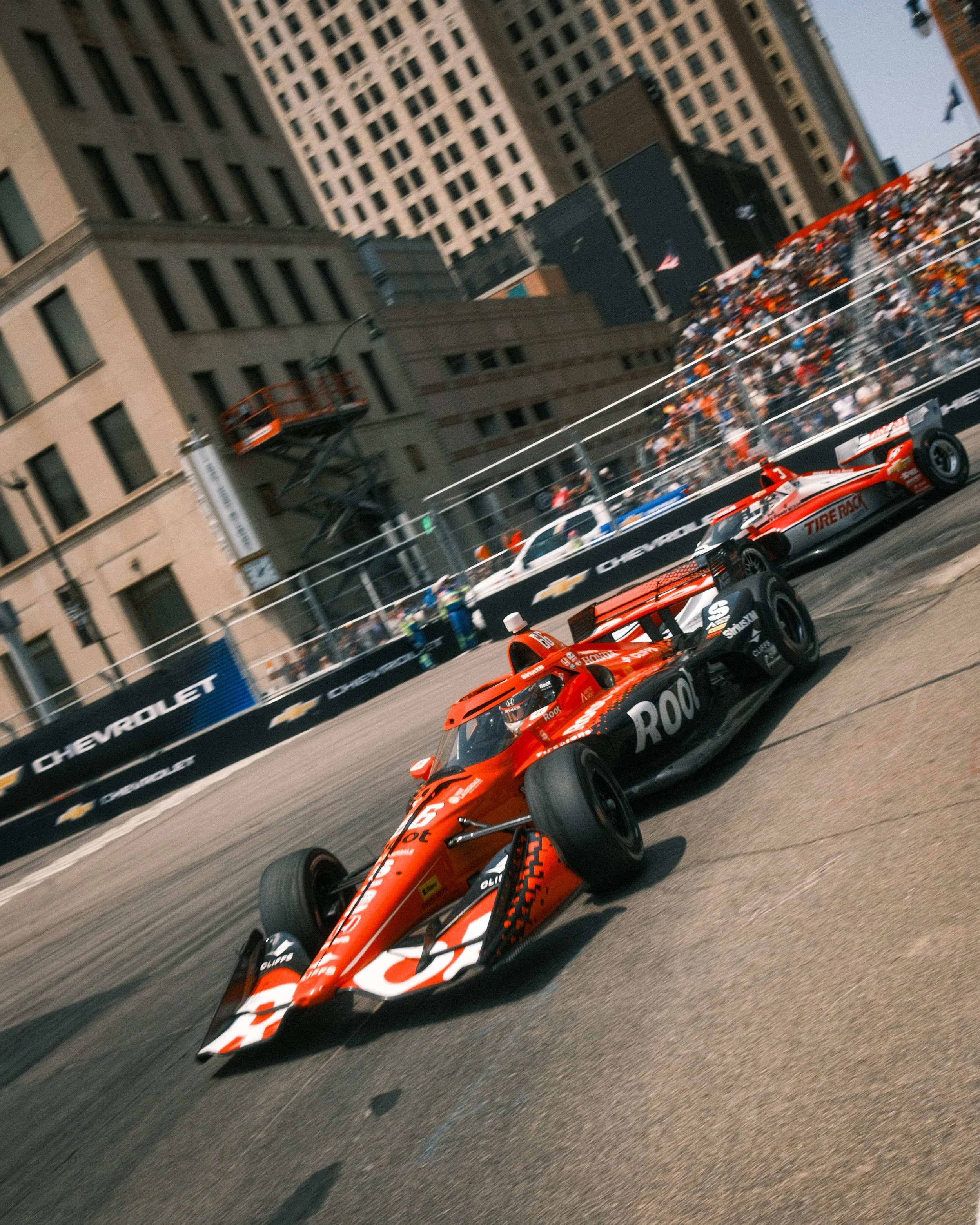 A red racing car on a city street circuit with a crowd of spectators behind a fence and tall buildings in the background.