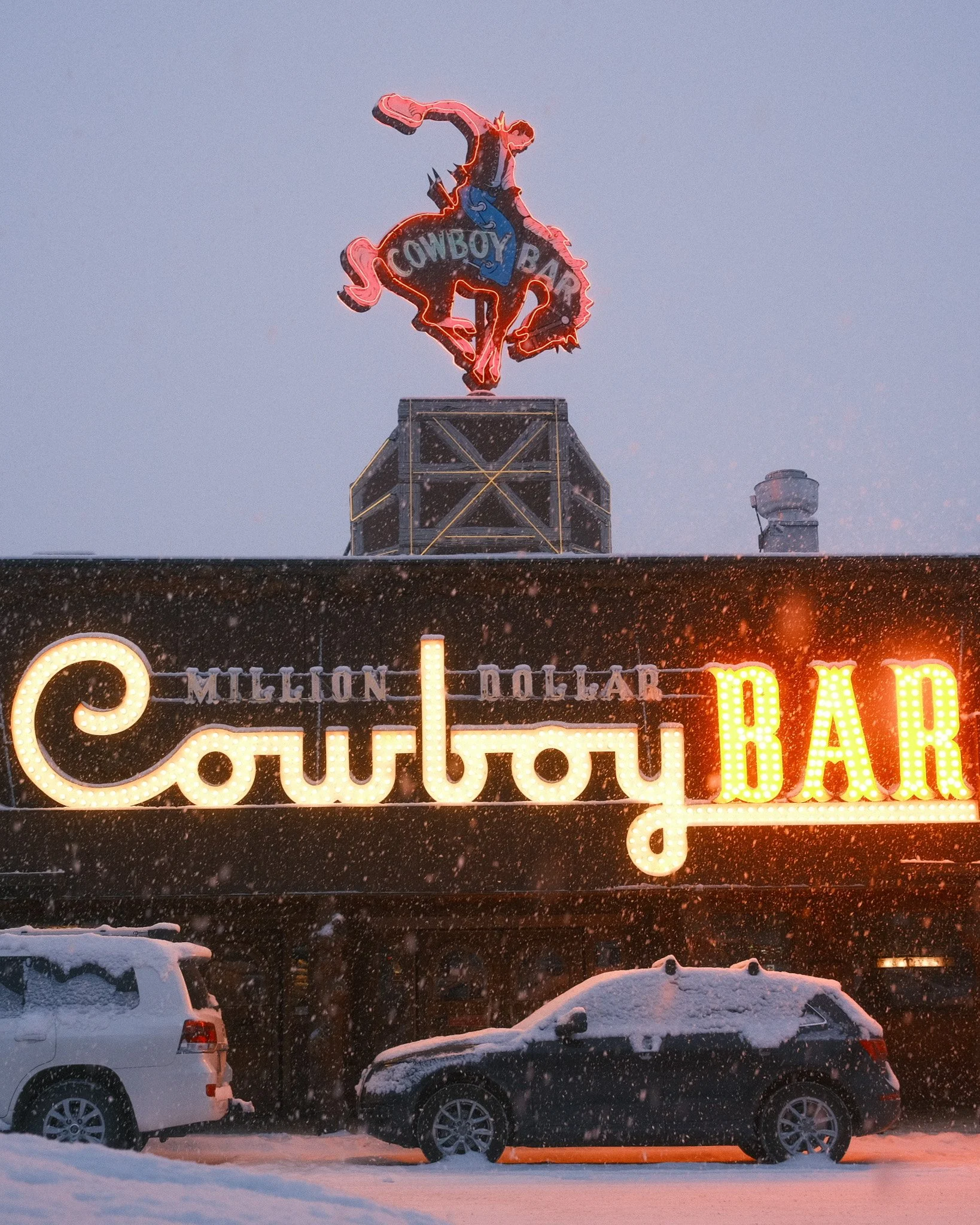 Neon sign for Cowboy Bar with cowboy riding a bucking horse, snowy weather, and parked cars in front.