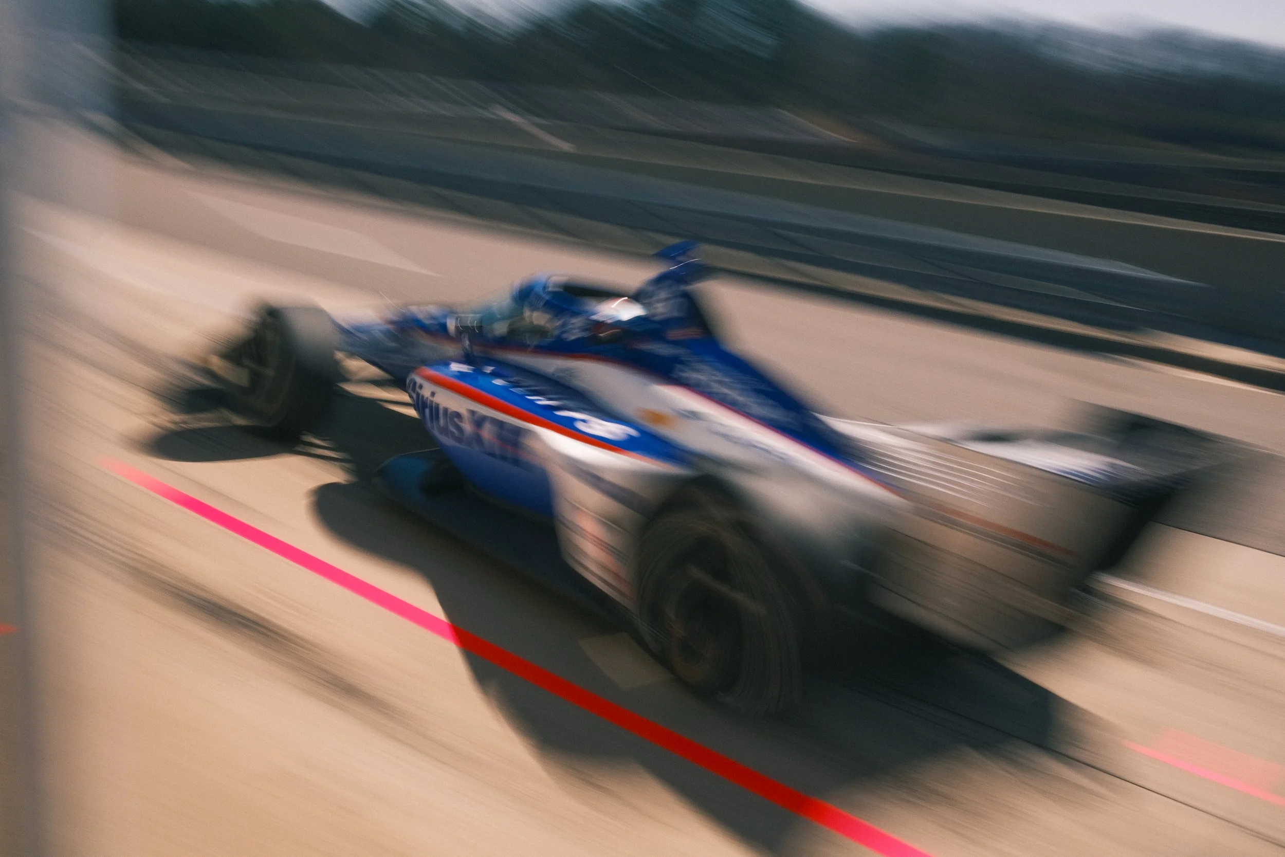 A blurred photo of a race car speeding on a race track, with a blue, white, and red color scheme and the word 'Aulus' visible on the side.