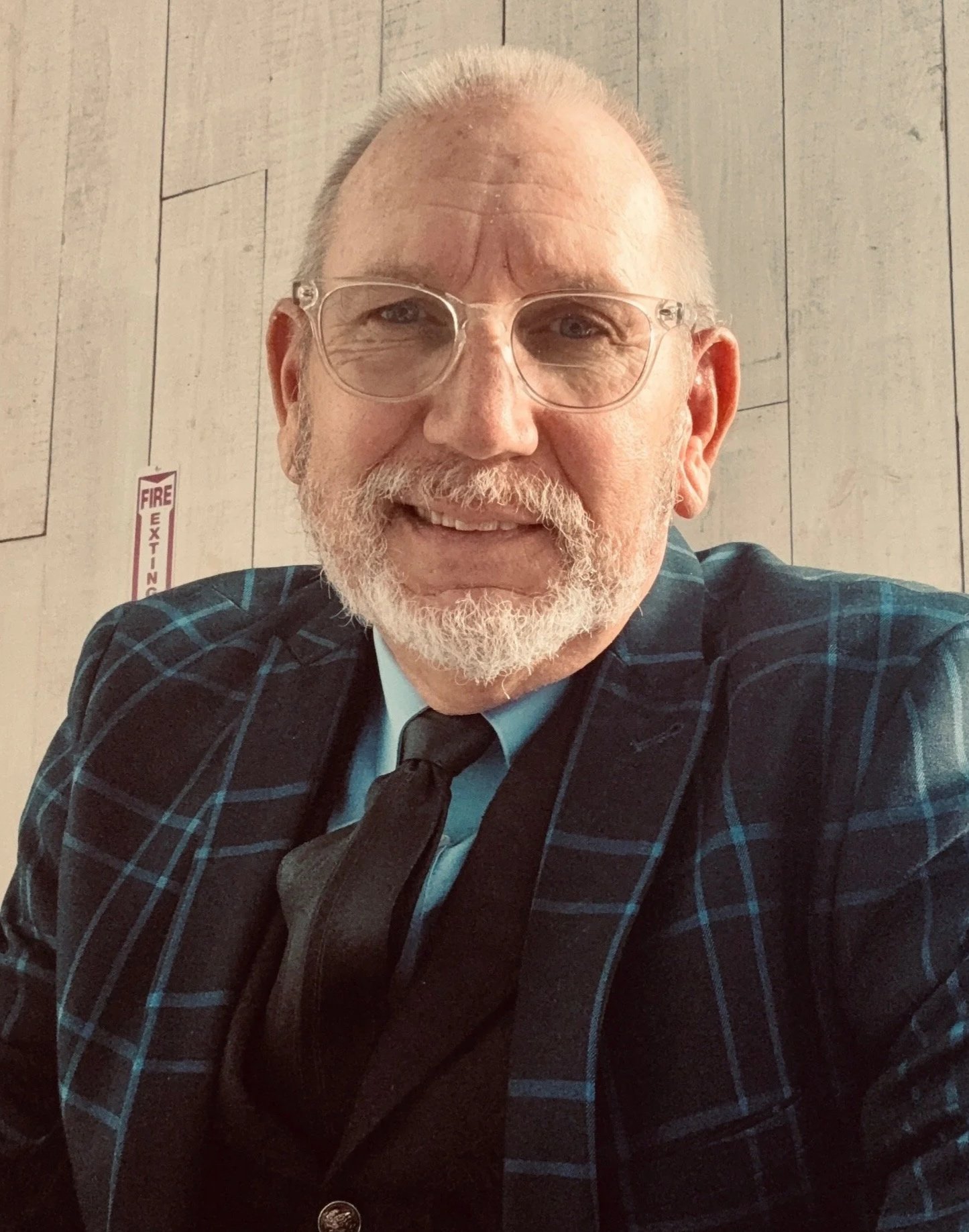 A man with glasses, a white beard, and a mustache smiling at the camera. He is wearing a blue and black plaid blazer, a light blue shirt, and a black tie. The background features wooden paneling and a fire exit sign.