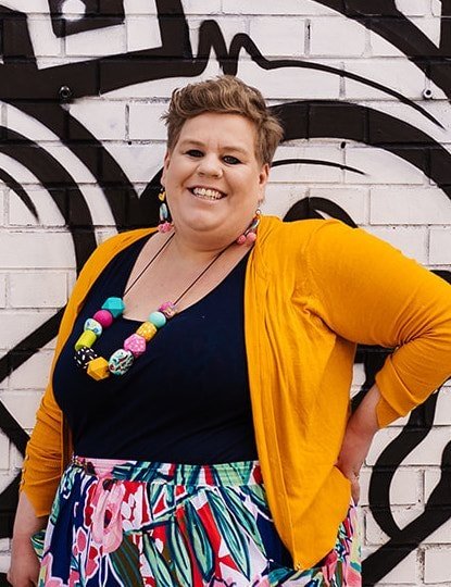 Meg smiling leaning against a graffitied brick wall.