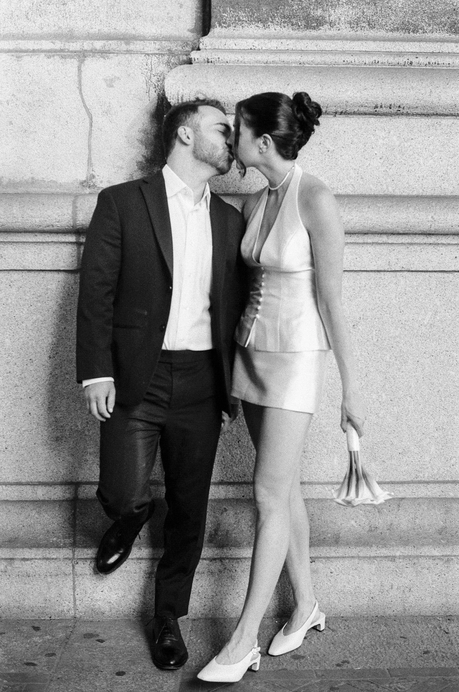 A black and white photo of a man in a suit and a woman in a dress holding hands and looking at each other, standing against a stone wall. The woman is holding a small bouquet of flowers.