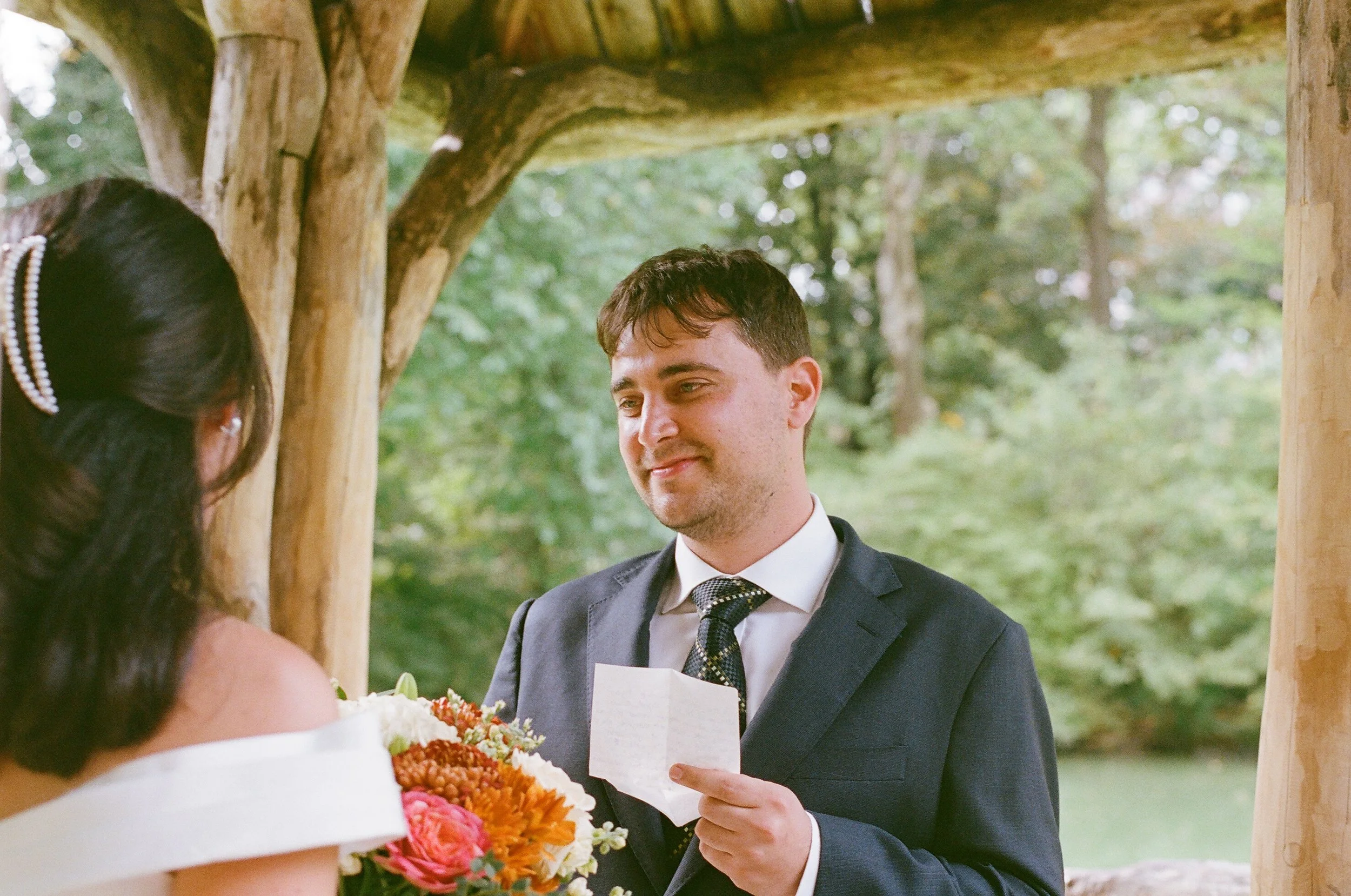 A woman in a white wedding gown is having a ring placed on her finger by a man in a suit during an outdoor wedding ceremony, with a officiant witnessing.