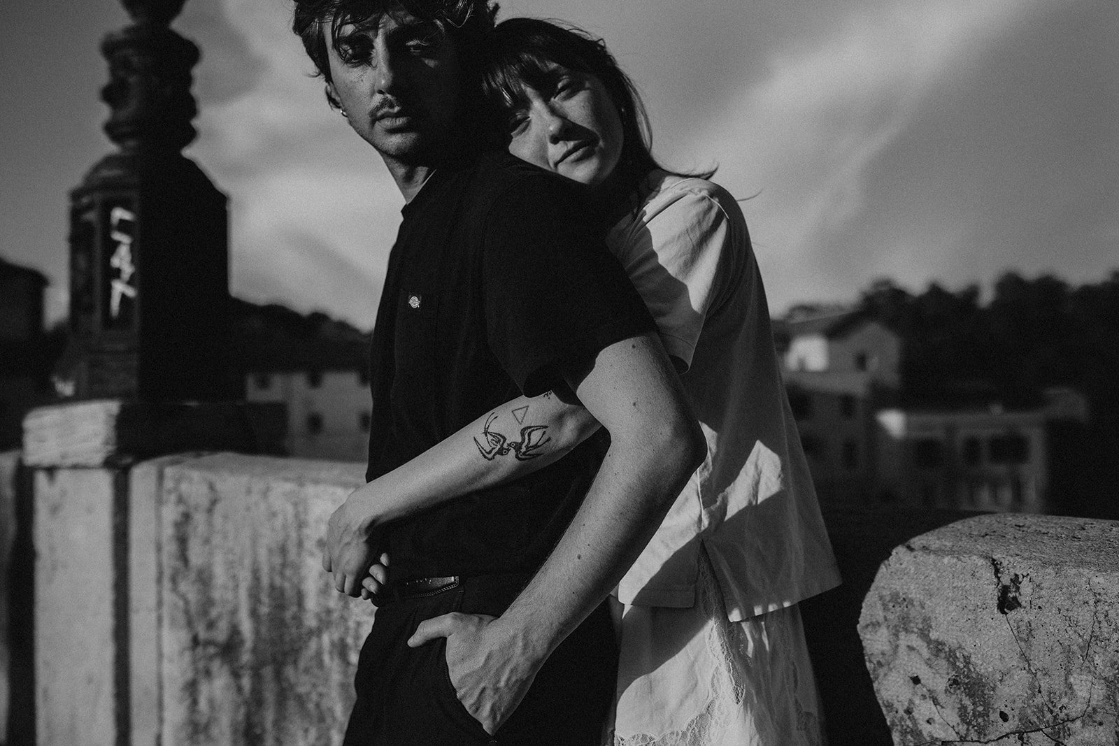 Black and white photo of a man and a woman leaning on a stone railing on a rooftop or terrace. The man has dark curly hair, a tattoo on his right forearm, and is wearing a dark shirt, looking at the camera. The woman has shoulder-length hair, is smiling slightly, and rests her head on the man's shoulder.