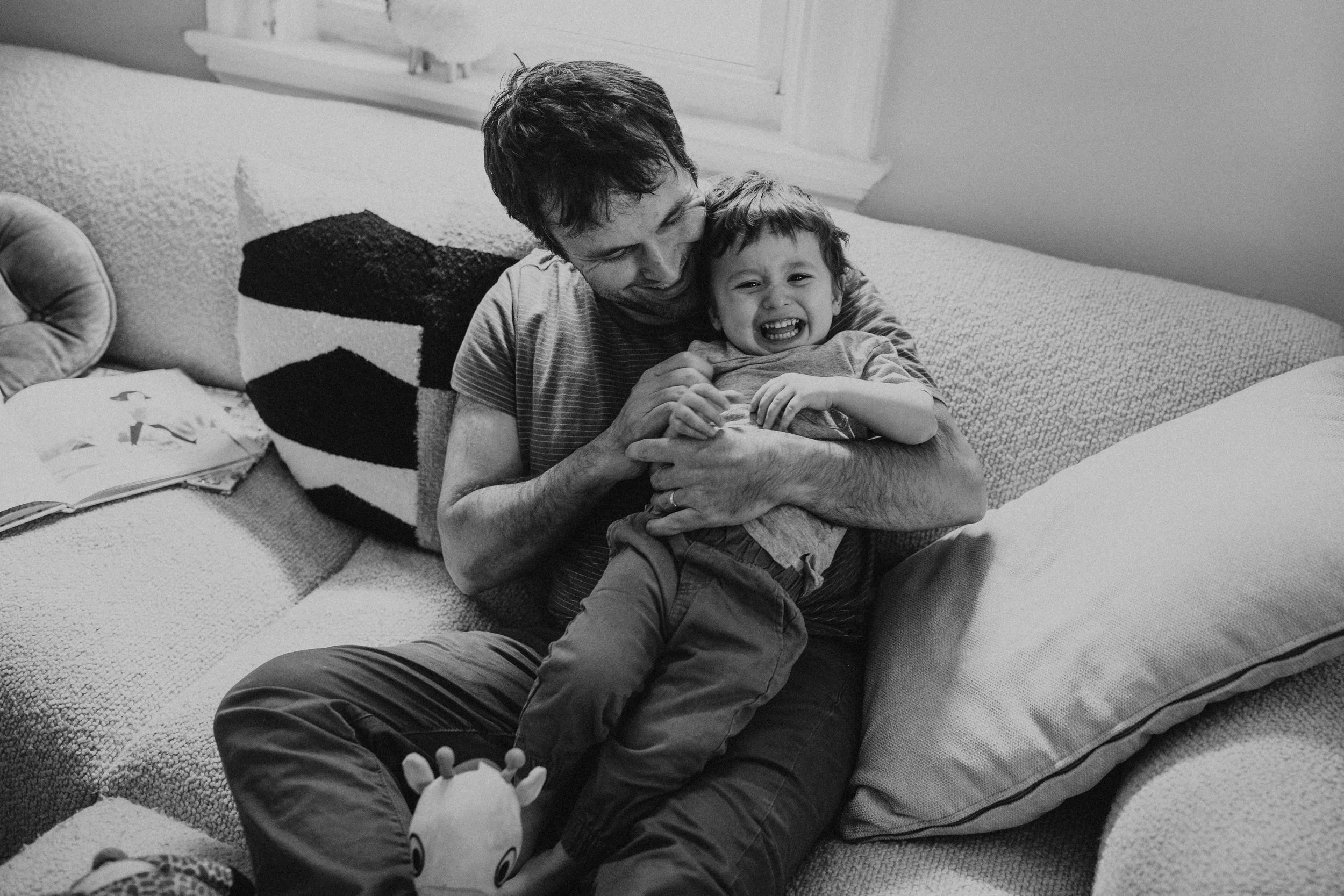 A man and a young boy sharing a joyful moment on a couch, with the boy laughing and the man smiling. The boy is sitting on the man's lap with their arms around each other.