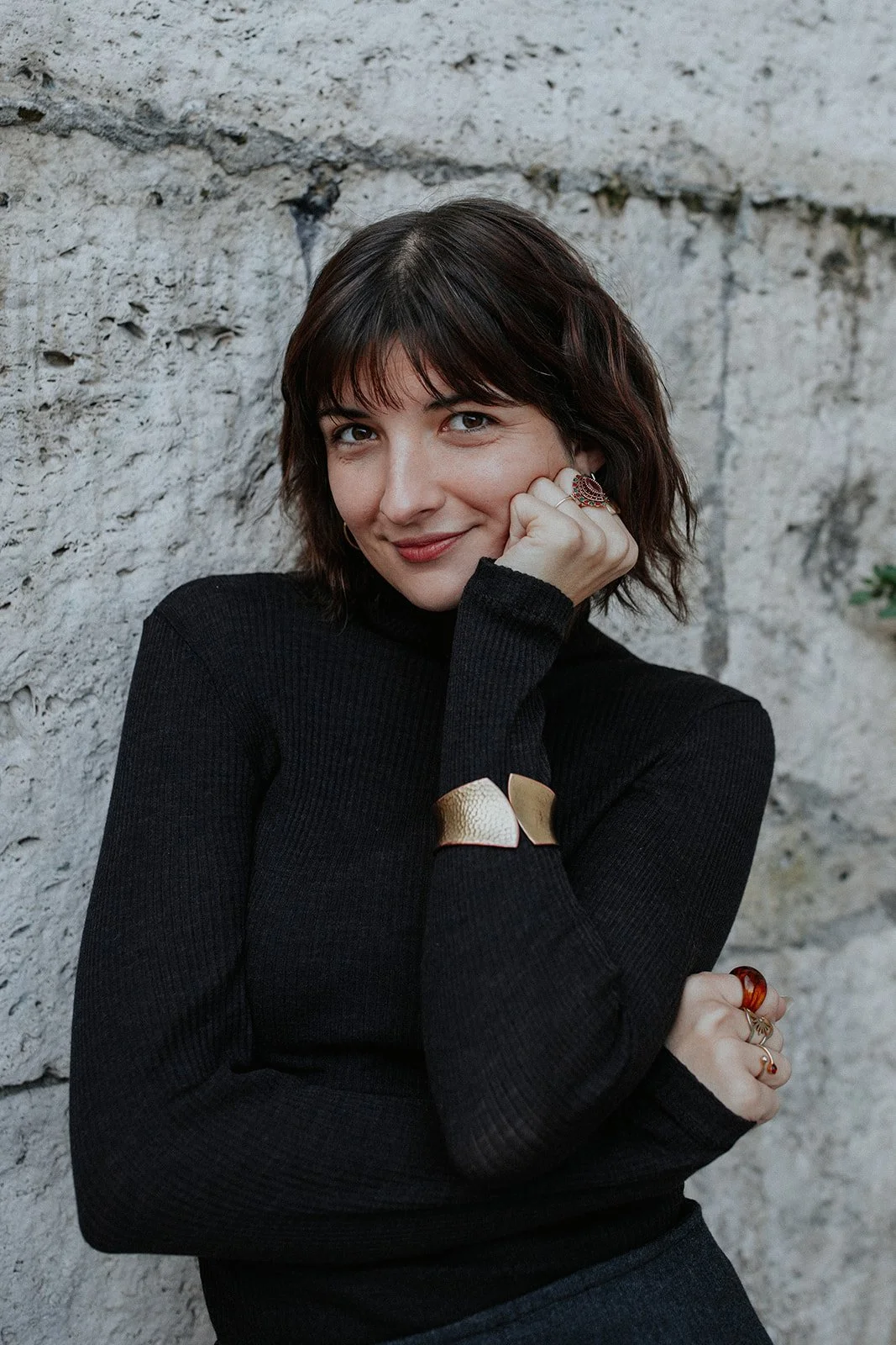 A woman with short dark hair wearing a black turtleneck and jewelry, smiling and resting her chin on her hand against a stone wall background.